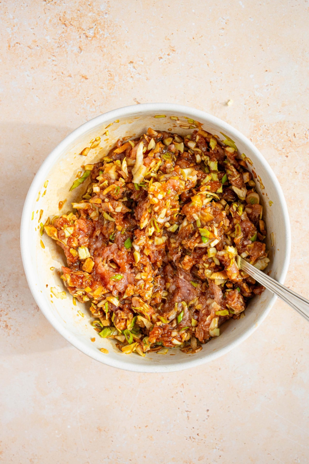 A large bowl with a spoon mixing a pork mixture for dumpling lasagna including ground pork, cabbage, green onion, mushrooms, garlic, ginger, sesame oil, and oyster sauce. The bowl is on a tan counter.