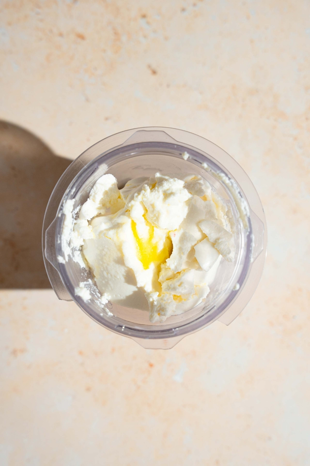 A glass blender with ricotta cheese and oil. The blender is on a white counter.