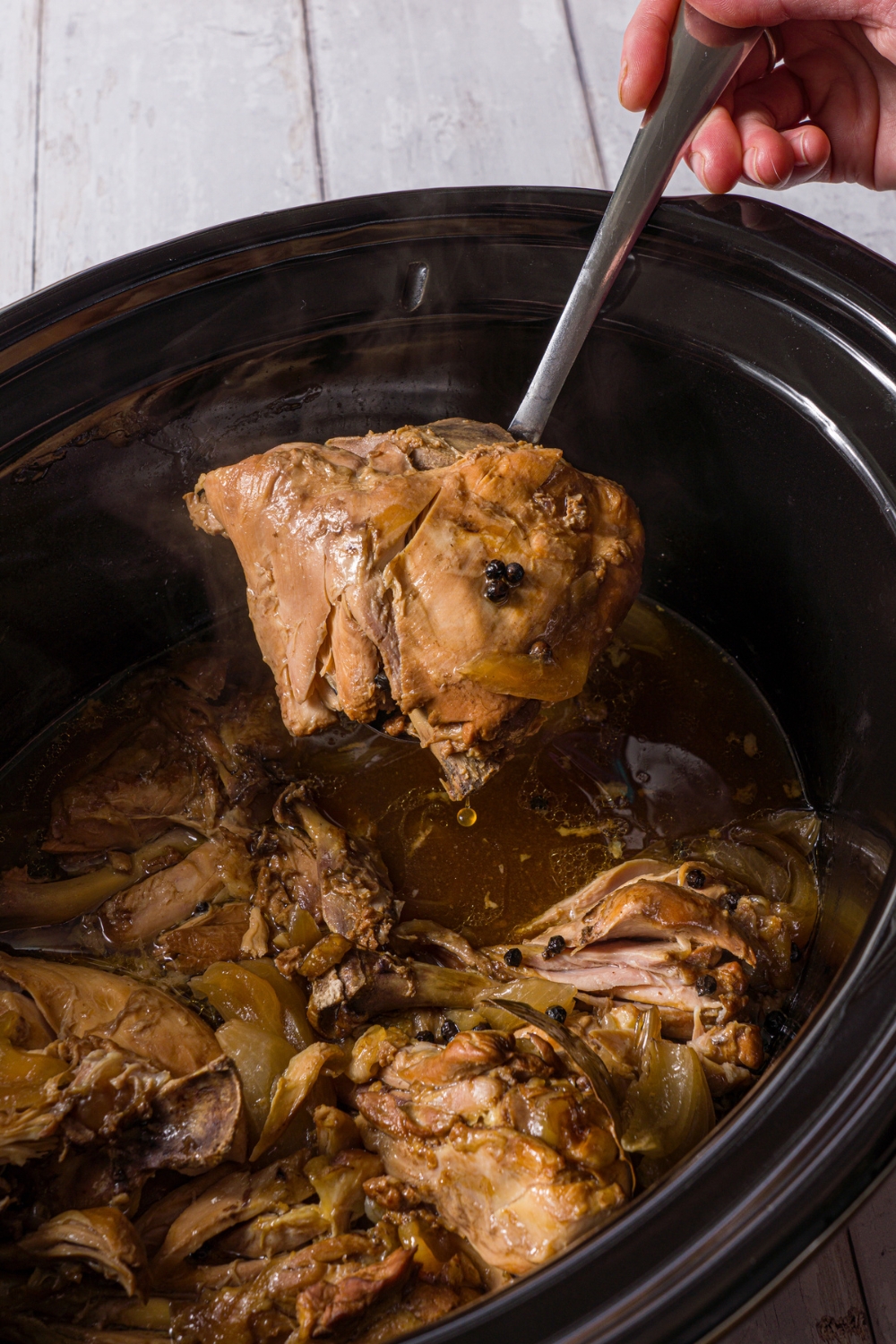 A crockpot with slow cooked chicken adobo. A spoon is removing chicken from the crockpot. The crockpot is on a wood counter.