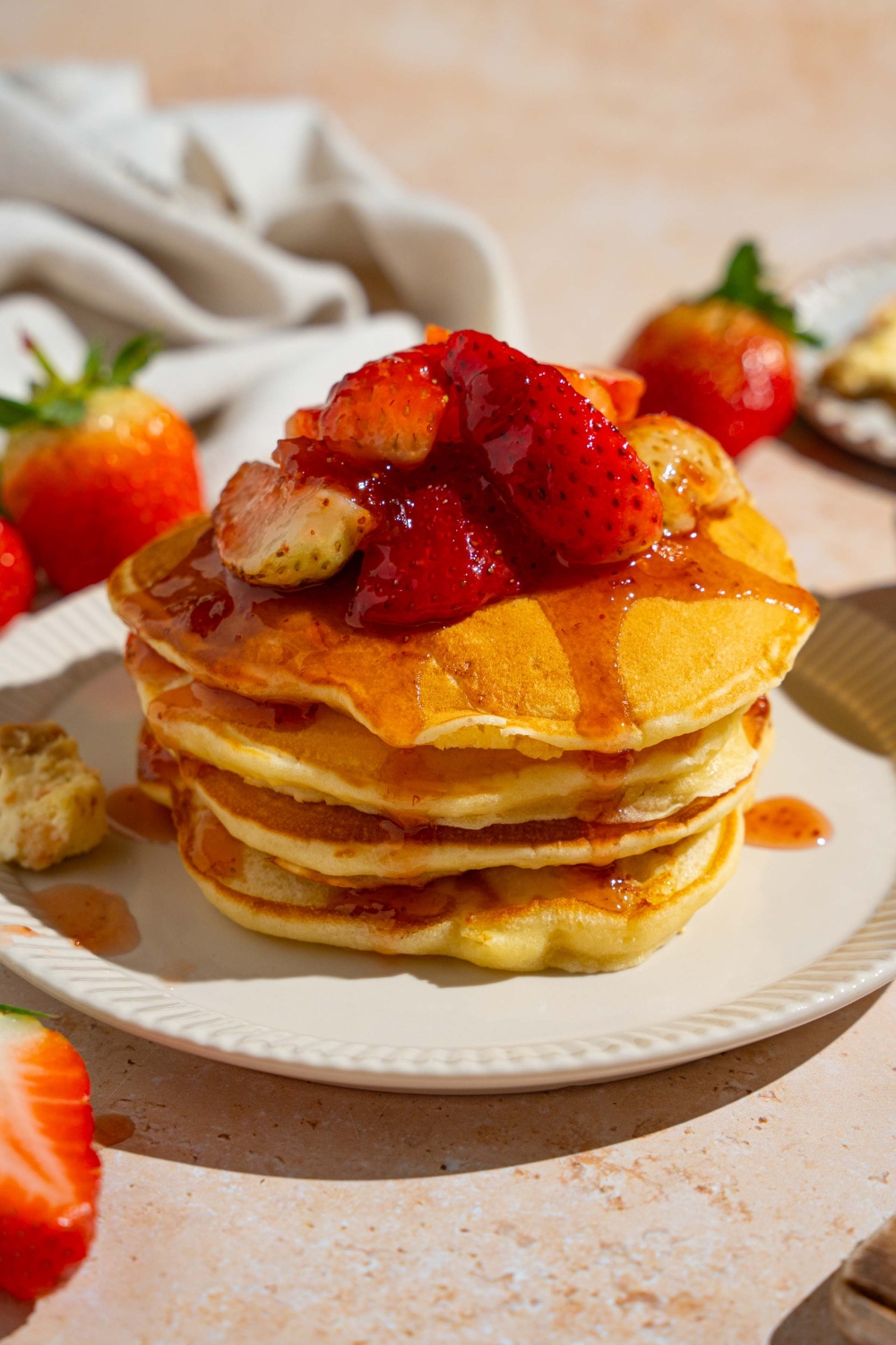 A white plate with a stack of IHOP New York cheesecake pancakes with a strawberry topping. The plate is on a tan counter with strawberries and a white cloth napkin.