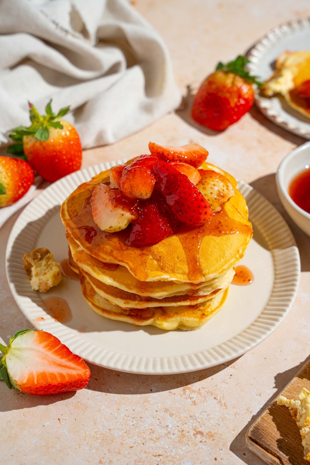 A white plate with a stack of IHOP New York cheesecake pancakes with a strawberry topping. The plate is on a tan counter with strawberries and a white cloth napkin.