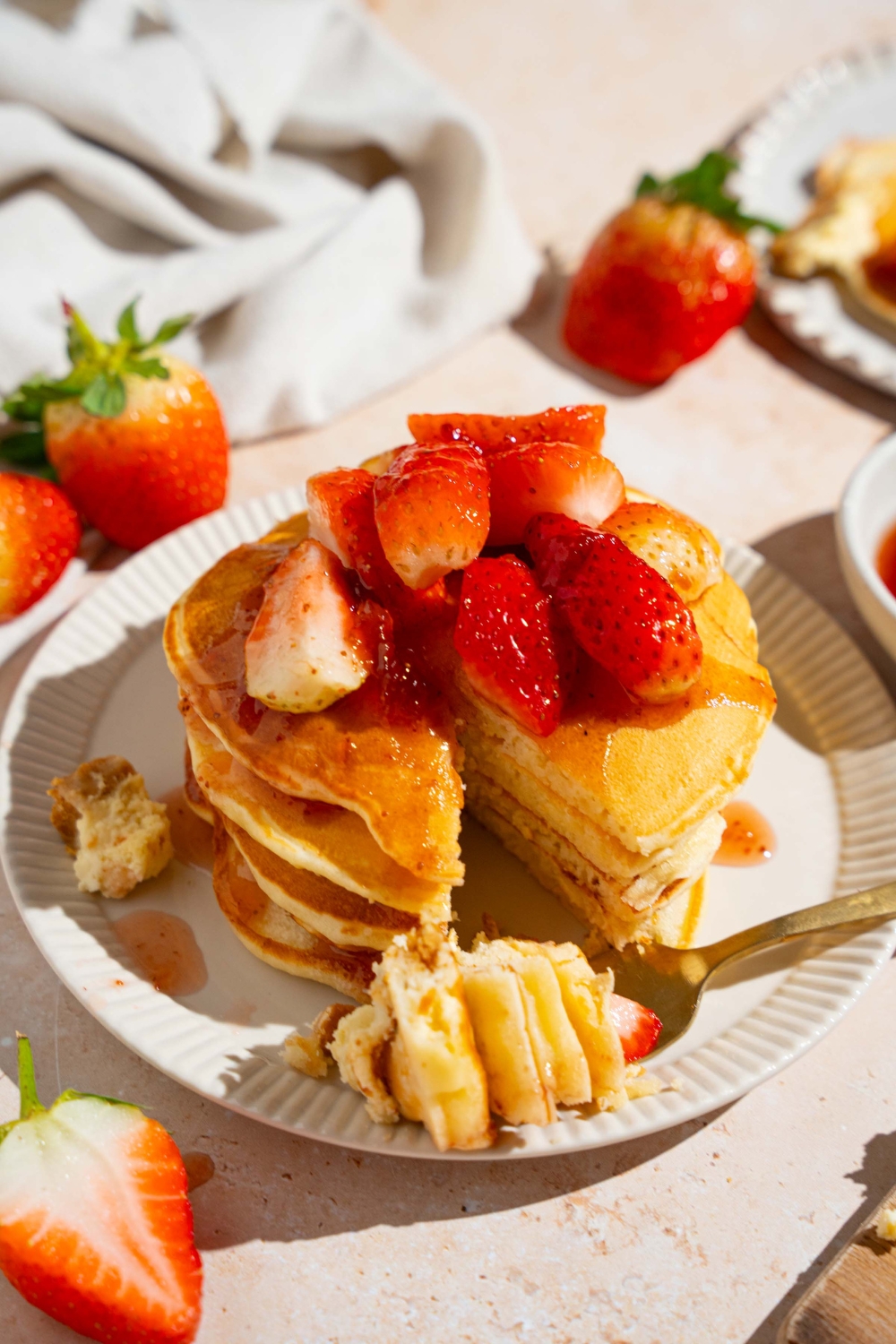 A white plate with a stack of IHOP New York cheesecake pancakes with a strawberry topping. A fork has removed a bite from the stack and is resting on the plate. The plate is on a tan counter with strawberries and a white cloth napkin.