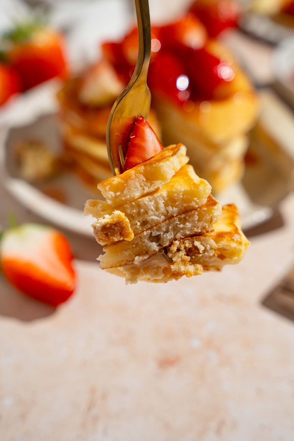 A close up of a fork with a bite of IHOP New York cheesecake pancakes. There is a plate of pancakes with a strawvberry topping on a tan counter blurred in the background.