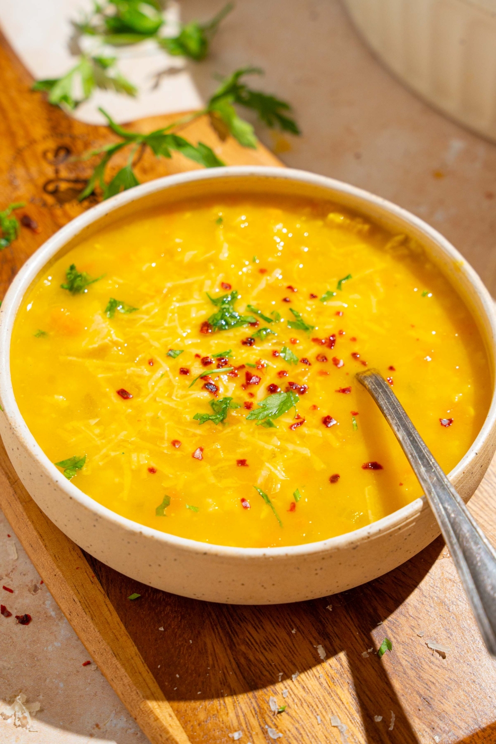 A white bowl of Italian penicillin soup garnished with fresh parsley and crushed red pepper. There is a spoon in the bowl. The bowl is on a wooden board on a white counter with fresh parsley.