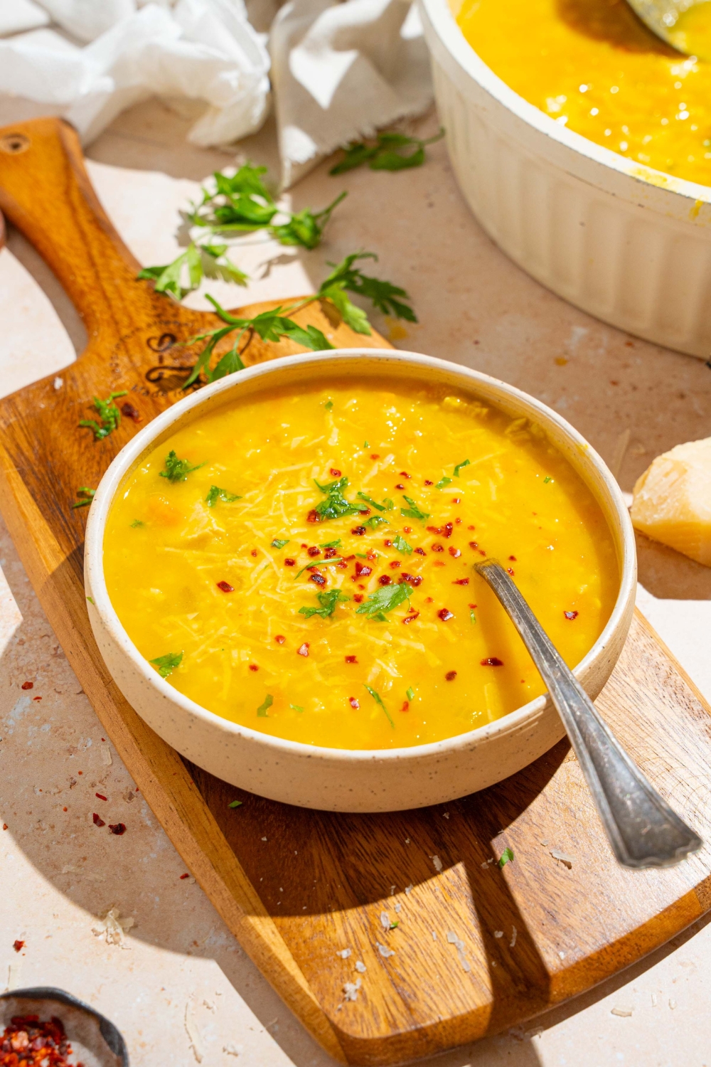 A white bowl of Italian penicillin soup garnished with fresh parsley and crushed red pepper. There is a spoon in the bowl. The bowl is on a wooden board on a white counter with a pot of soup and fresh parsley.
