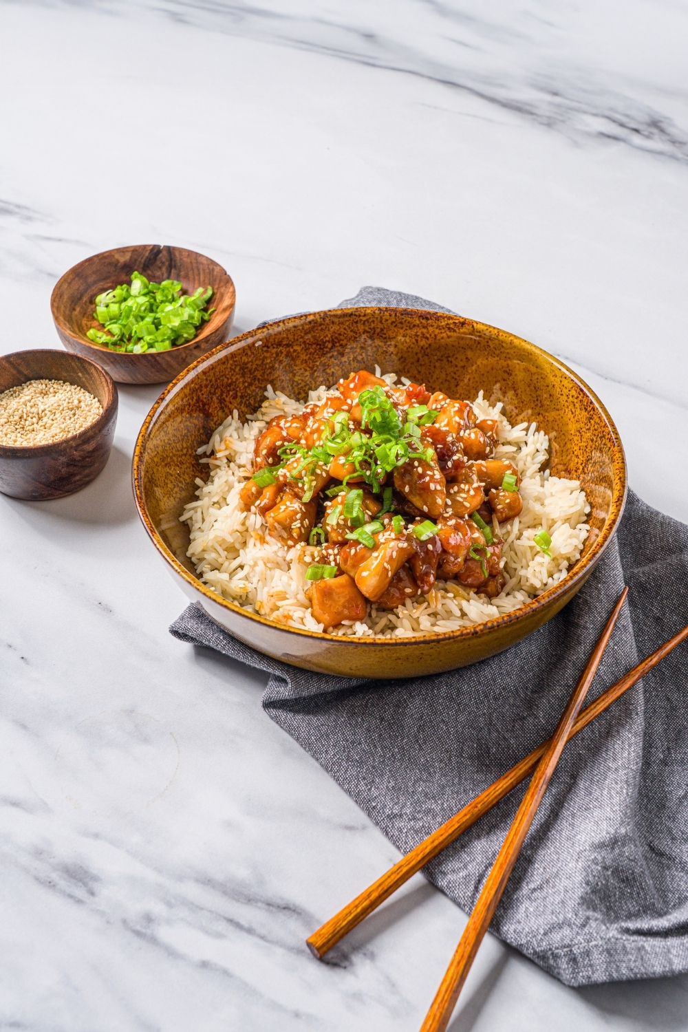 A bowl of Korean sticky chicken served over rice garnished with sliced green onions and sesame seeds. The bowl is on a blue cloth napkin with a pair of chopsticks on a marble counter with small bowls of garnishes.