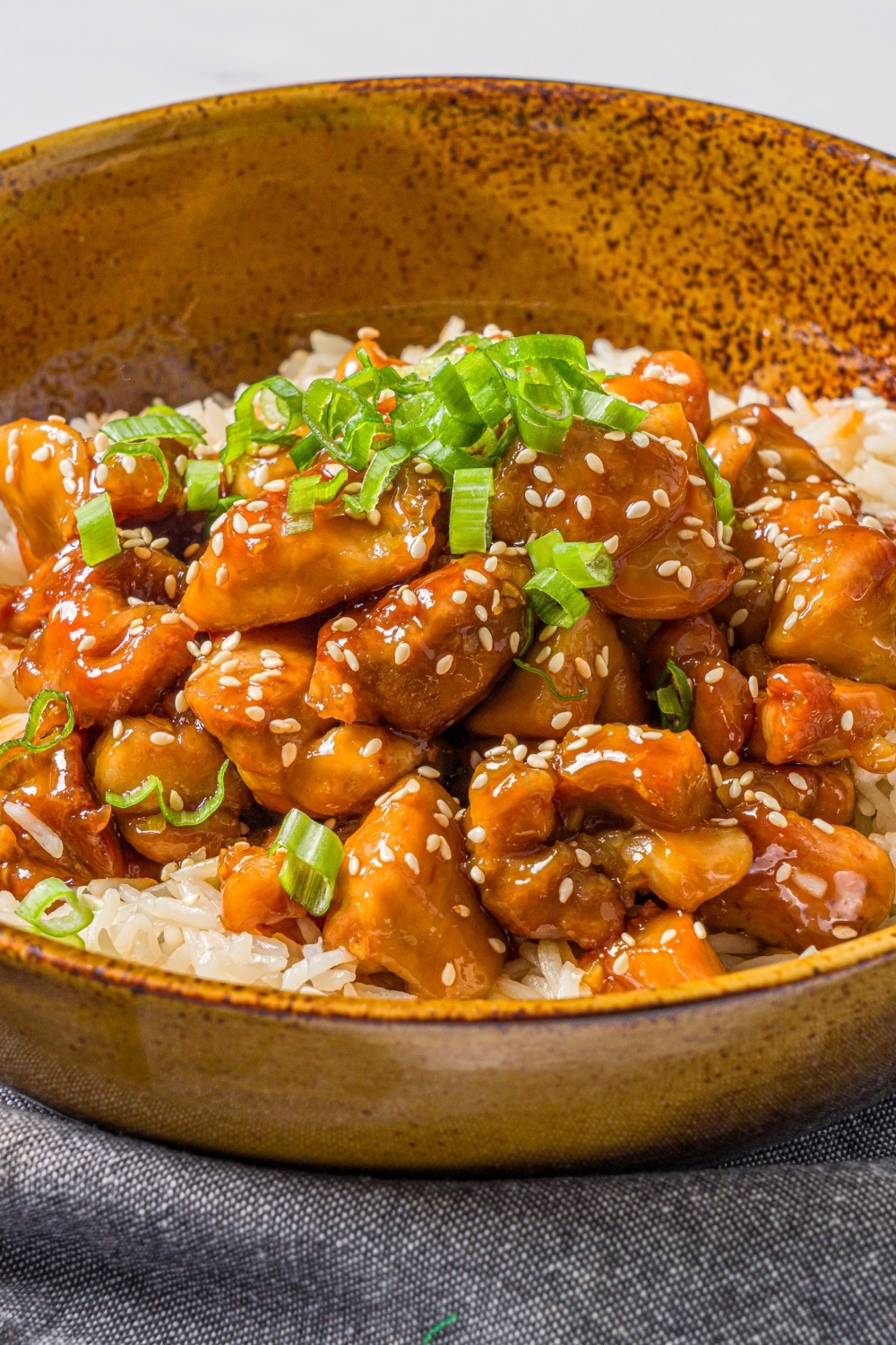 A bowl of Korean sticky chicken served over rice garnished with sliced green onions and sesame seeds. The bowl is on a blue cloth napkin on a marble counter.