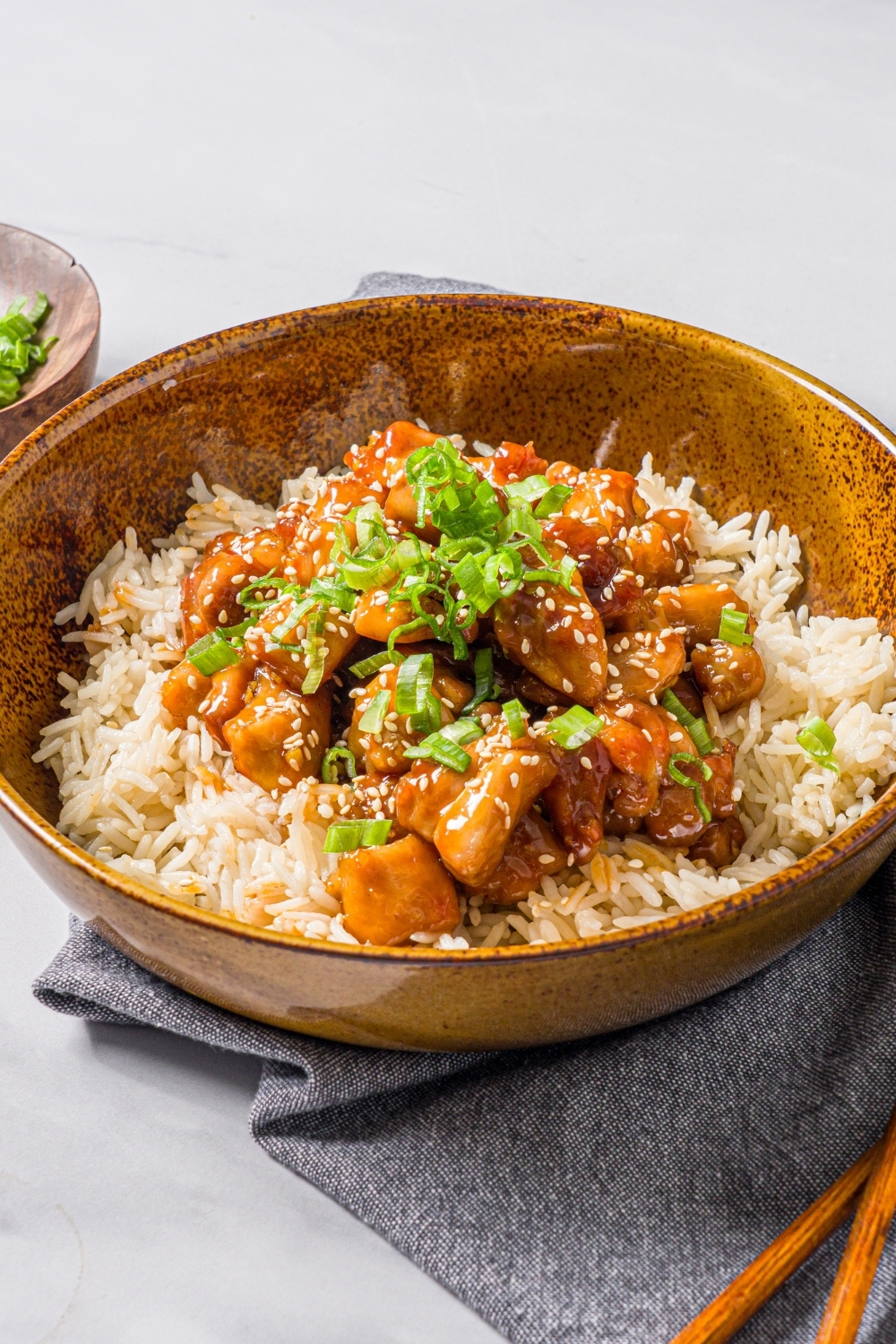 A bowl of Korean sticky chicken served over rice garnished with sliced green onions and sesame seeds. The bowl is on a blue cloth napkin with a pair of chopsticks on a marble counter.