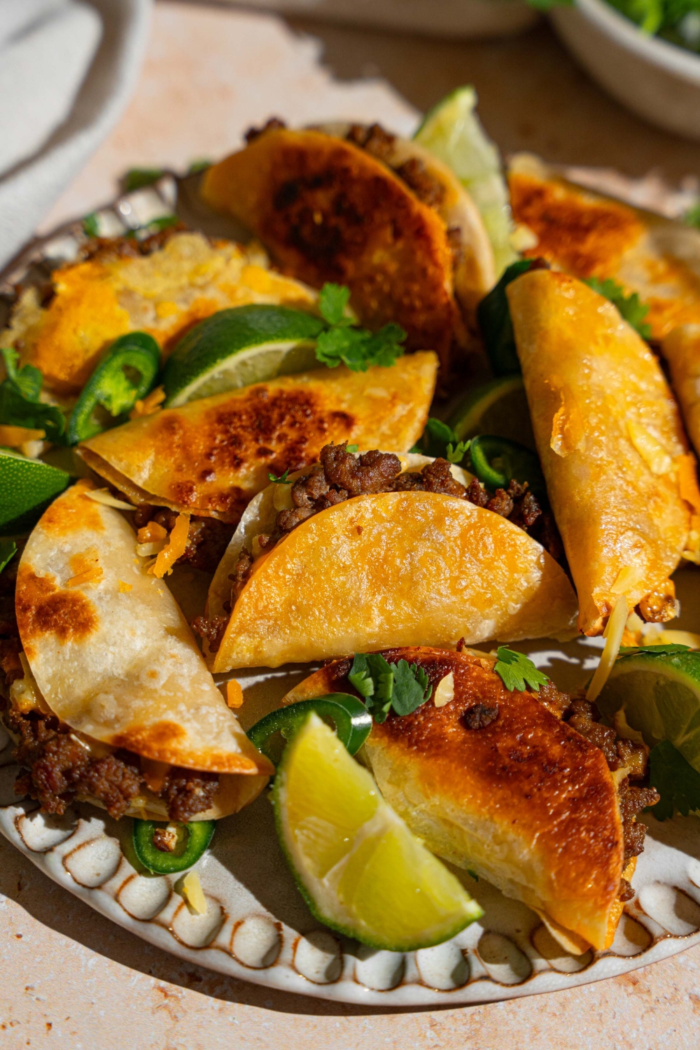 A plate of baked mini beef tacos garnished with cilantro, sliced jalapeño, and lime wedges. The plate is on a tan counter.
