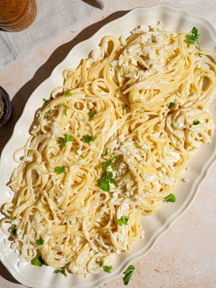 A white platter with Mexican white spaghetti tossed with crumbled queso fresco and garnished with fresh parsley. The platter is on a tan counter.