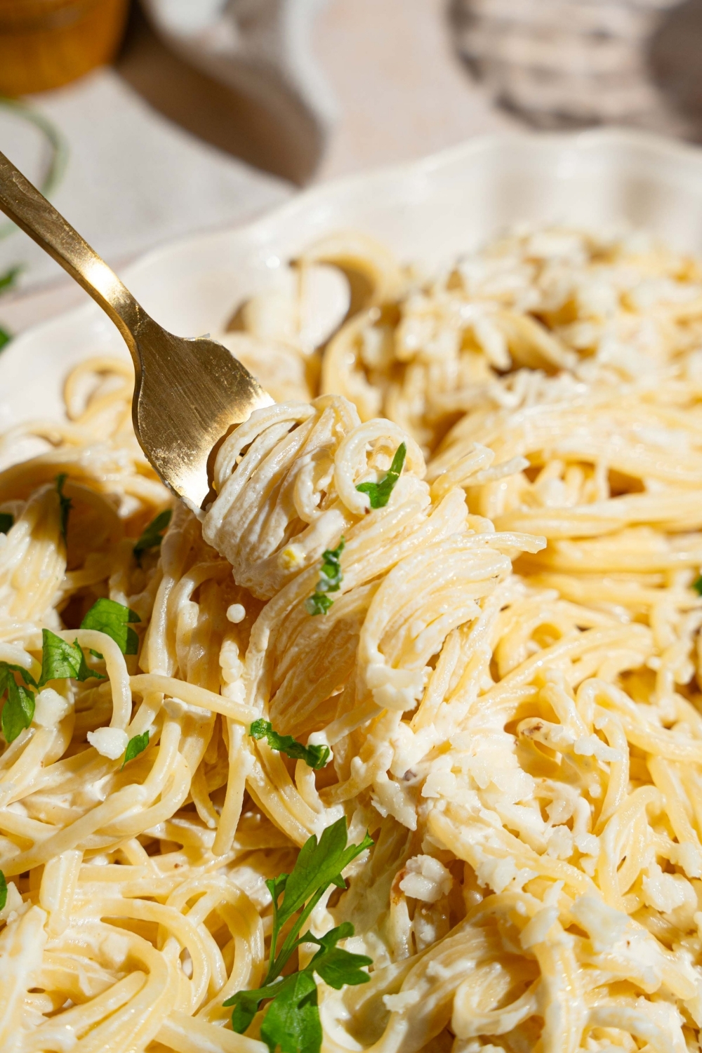 A white platter with Mexican white spaghetti tossed with crumbled queso fresco and garnished with fresh parsley. A fork is twirling spaghetti from the platter.