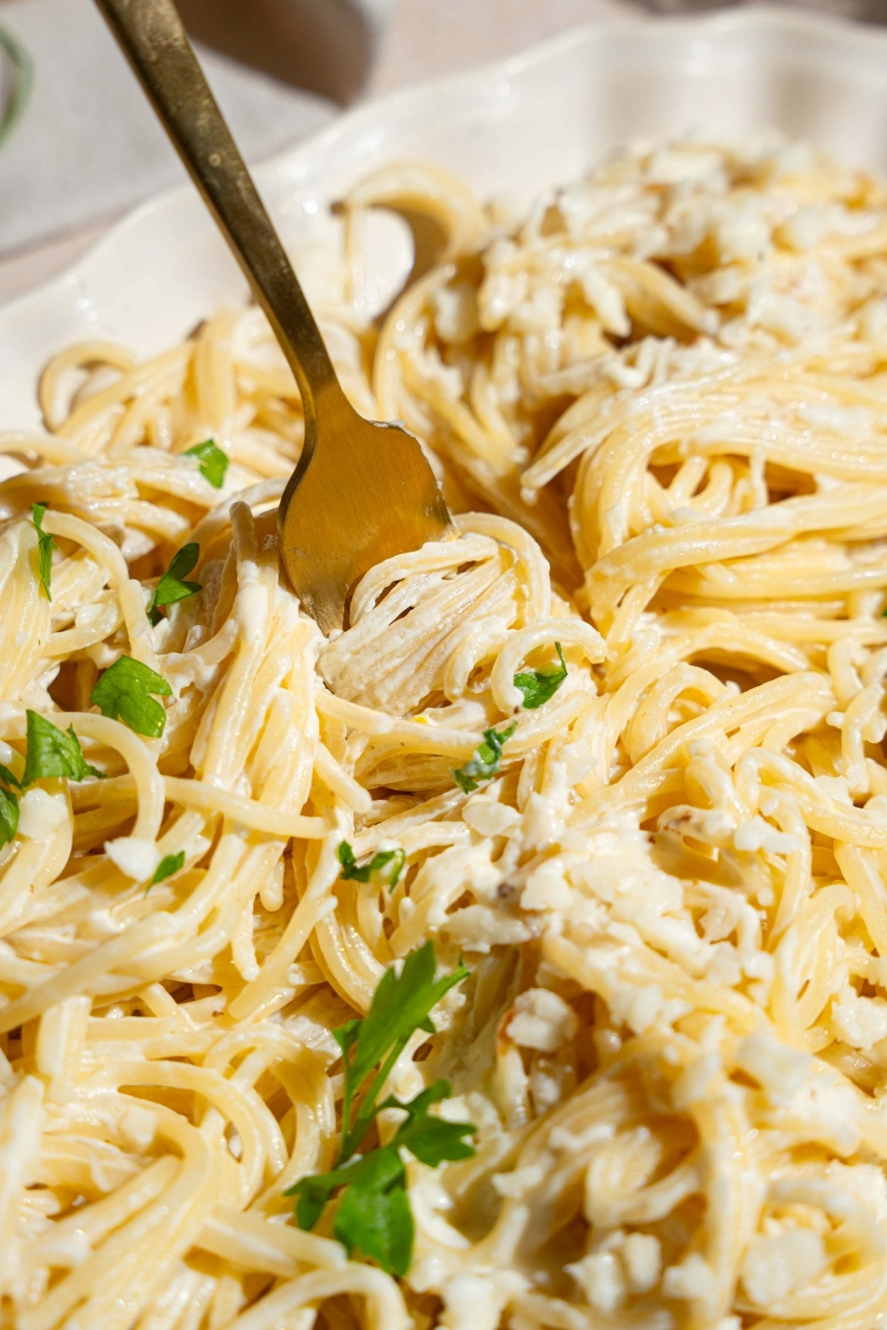 A white platter with Mexican white spaghetti tossed with crumbled queso fresco and garnished with fresh parsley. A fork is twirling spaghetti from the platter.