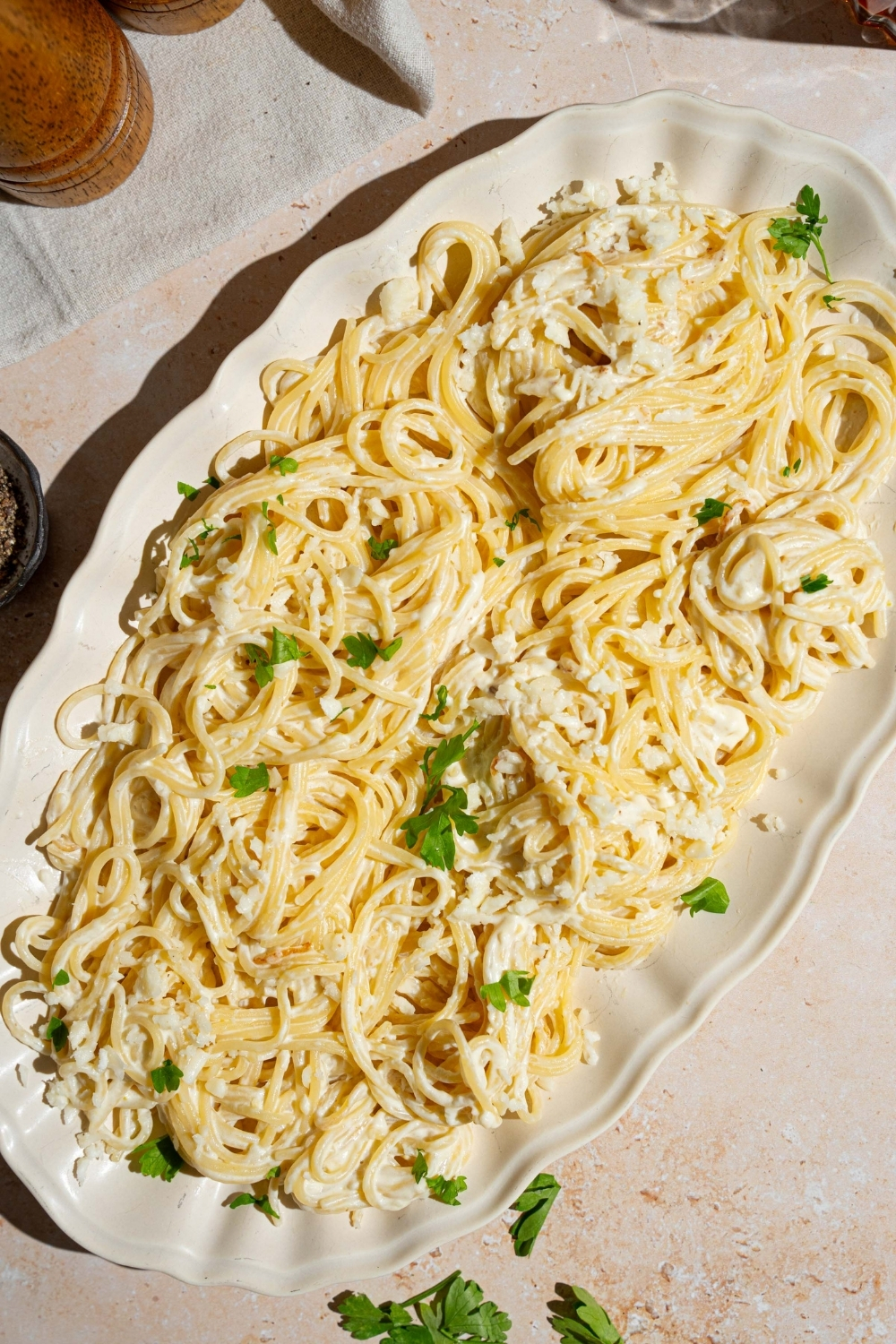 A white platter with Mexican white spaghetti tossed with crumbled queso fresco and garnished with fresh parsley. The platter is on a tan counter.