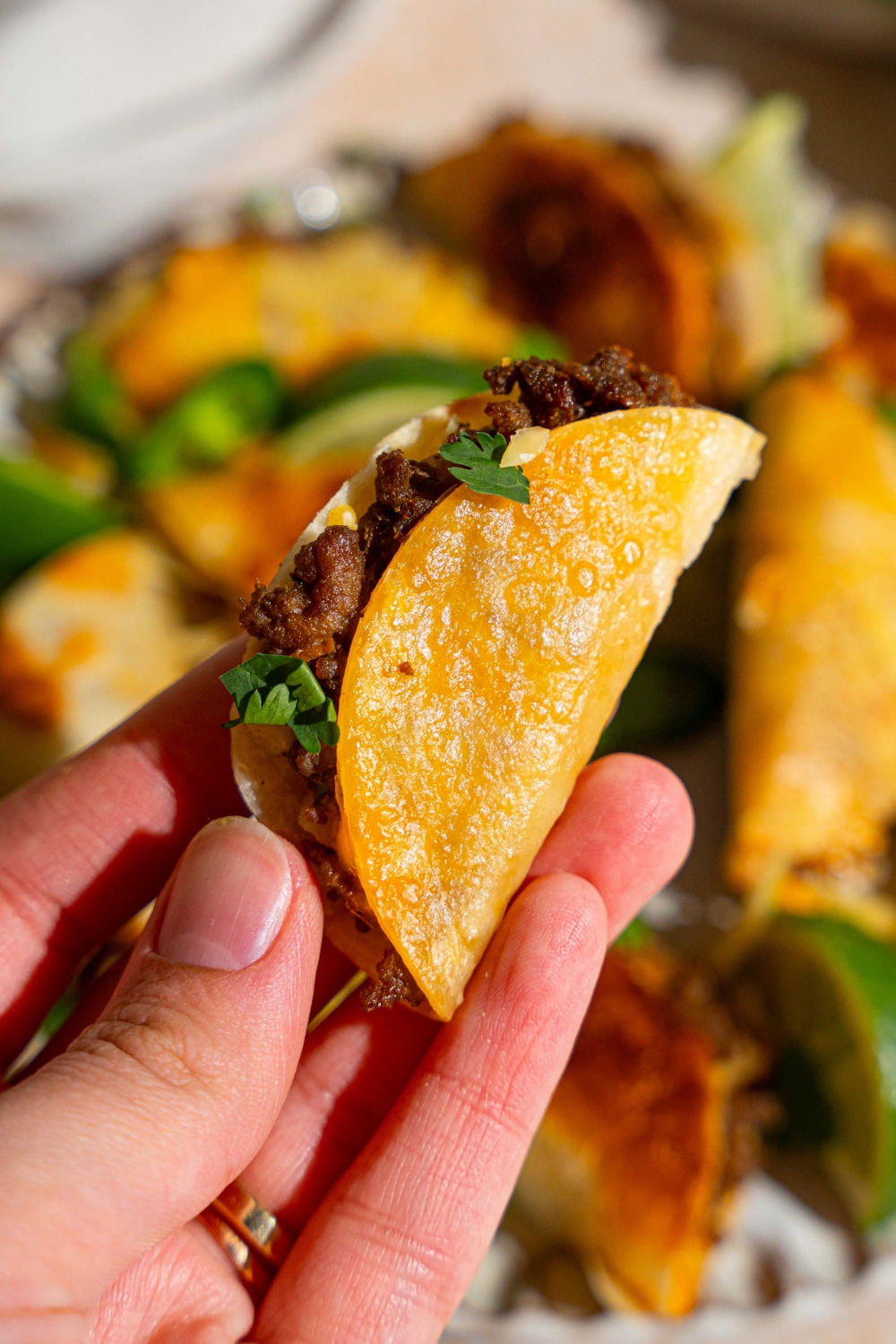 A close up of a hand holding a mini beef taco garnished with fresh cilantro with a plate of tacos blurred in the background.