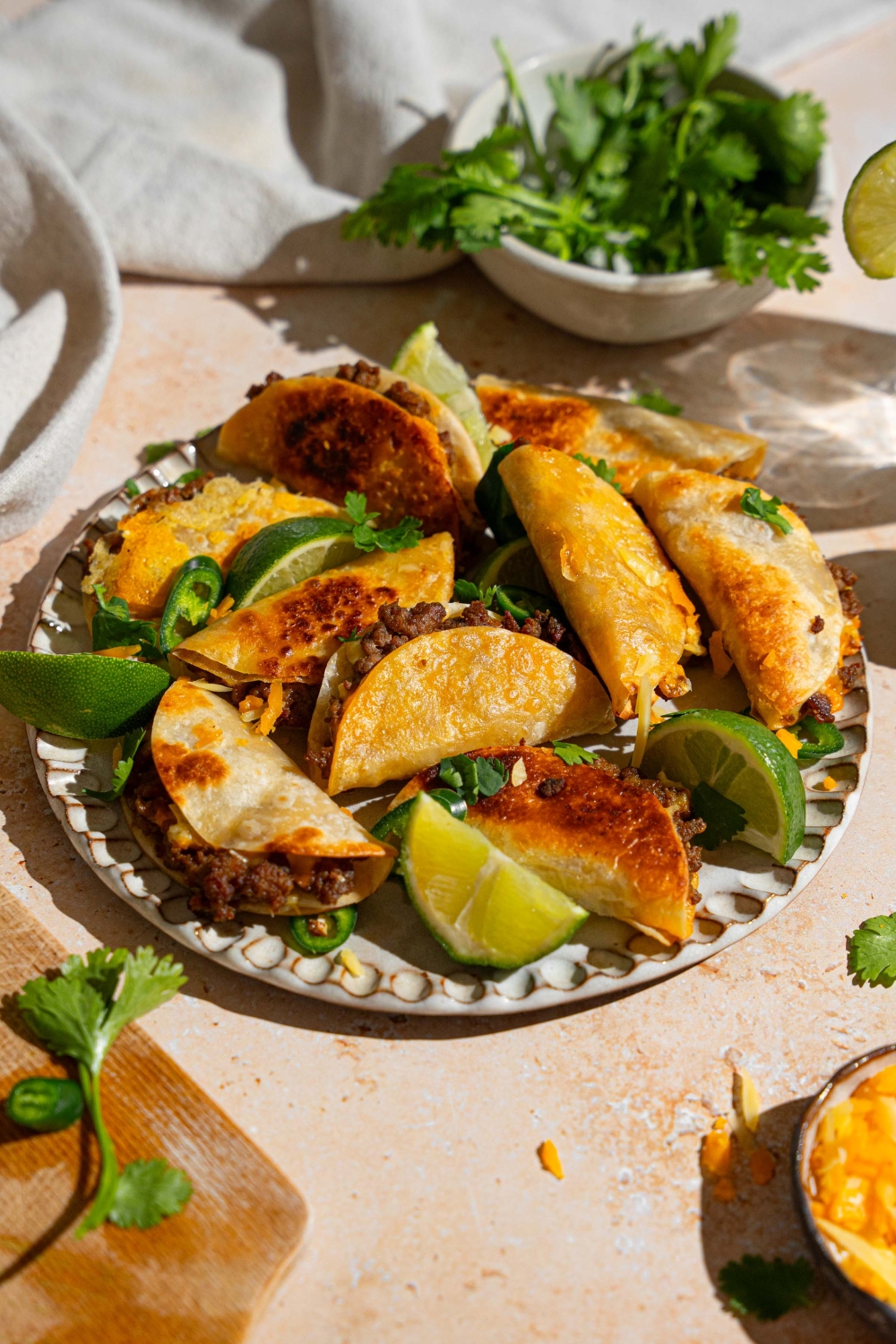 A plate of baked mini beef tacos garnished with cilantro, sliced jalapeño, and lime wedges. The plate is on a tan counter with a bowl of cilantro and white cloth napkin.