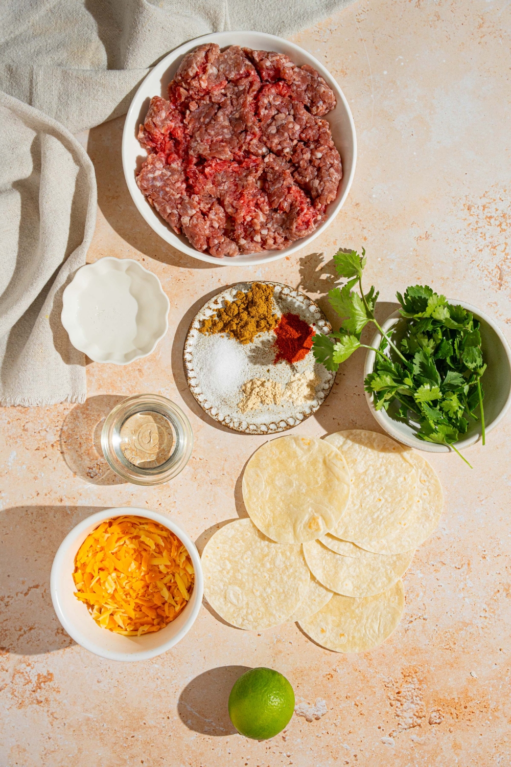 An overhead shot of several bowls in various sizes containing ingredients to make mini beef tacos including ground beef, mini tortilla shells, shredded cheese, cilantro, and seasonings.