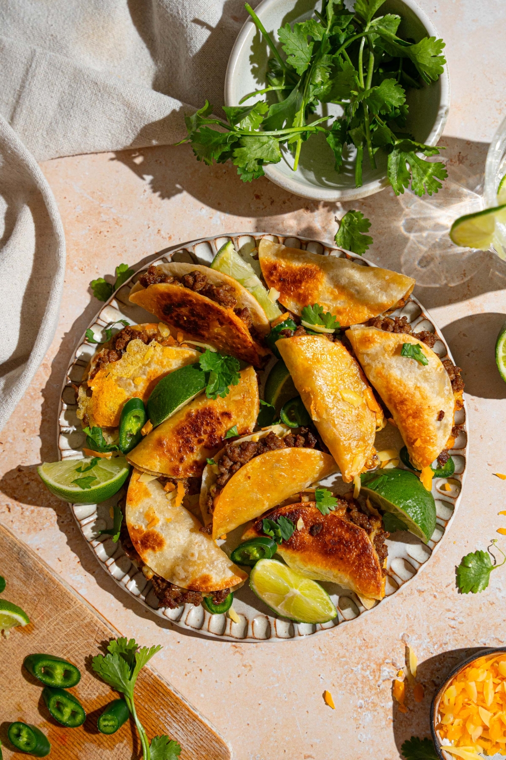 A plate of baked mini beef tacos garnished with cilantro, sliced jalapeño, and lime wedges. The plate is on a tan counter with a bowl of cilantro and white cloth napkin.