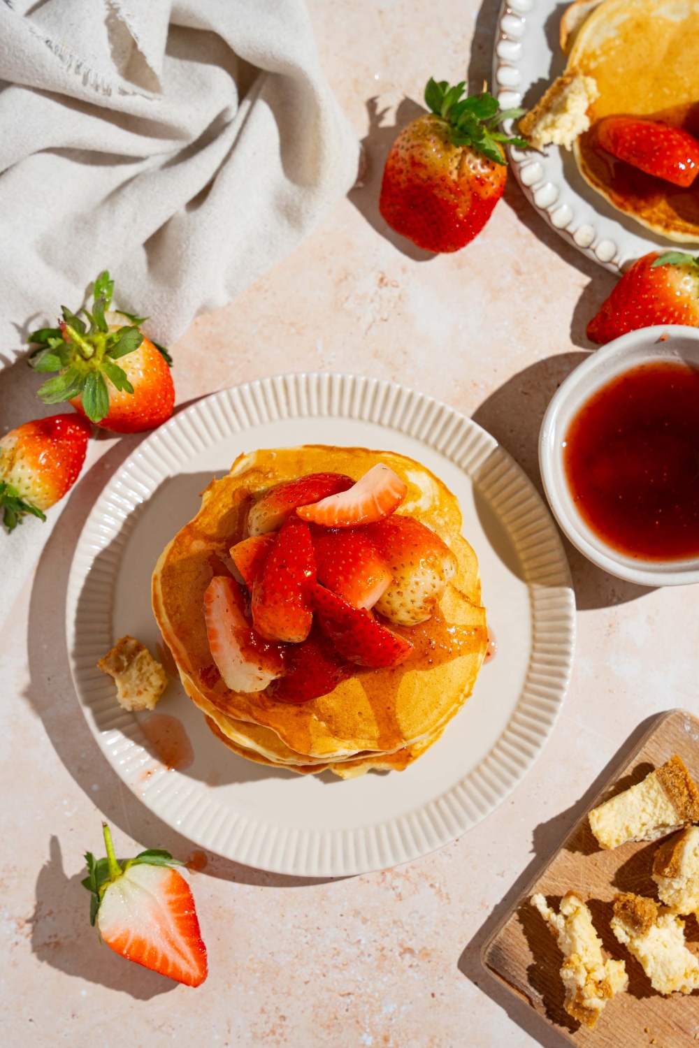 A white plate with a stack of IHOP New York cheesecake pancakes with a strawberry topping. The plate is on a tan counter with strawberries and a white cloth napkin.