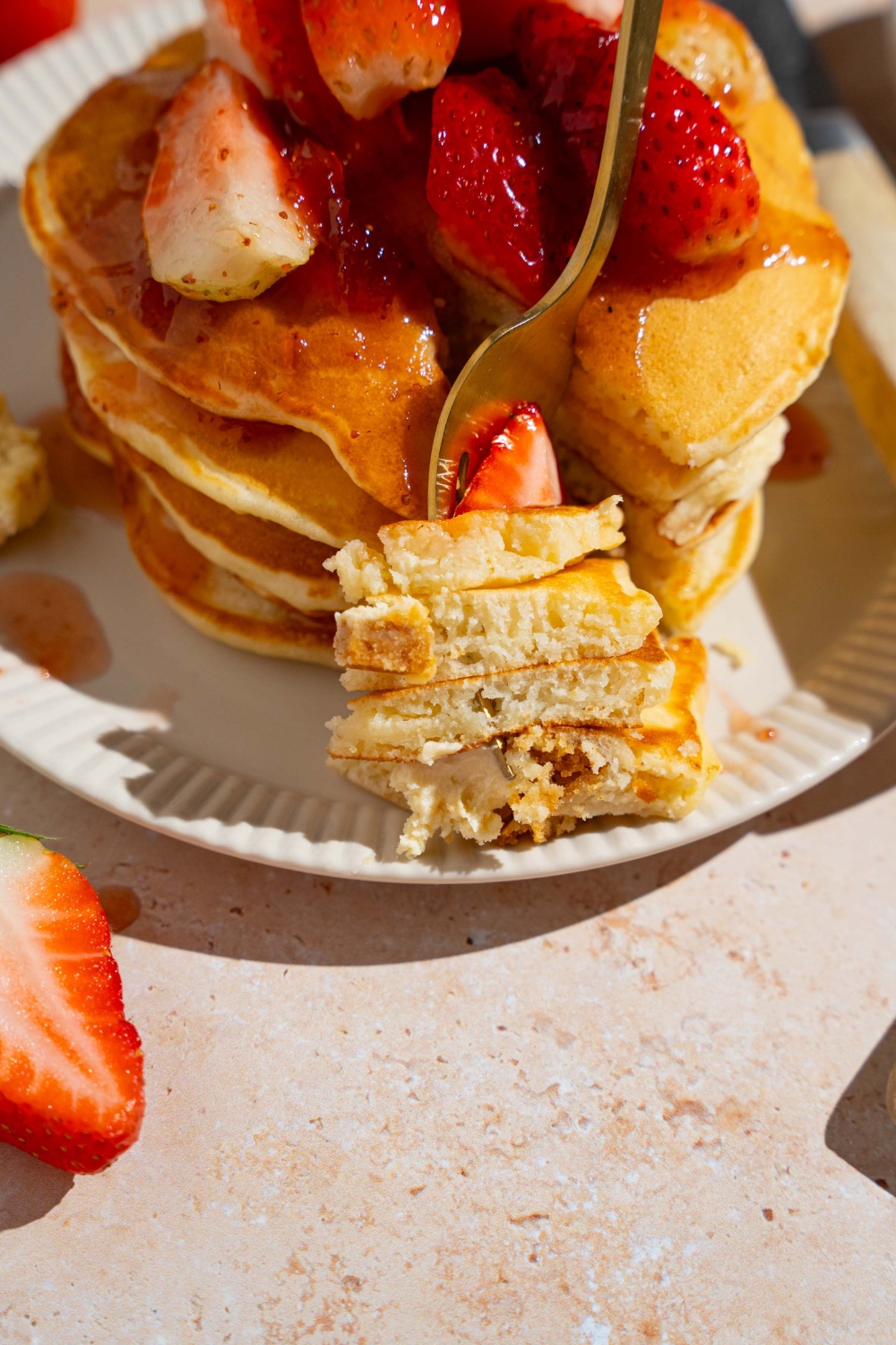 A white plate with a stack of IHOP New York cheesecake pancakes with a strawberry topping. A fork has taken a bite from the stack and is resting on the plate. The plate is on a tan counter with strawberries and a white cloth napkin.