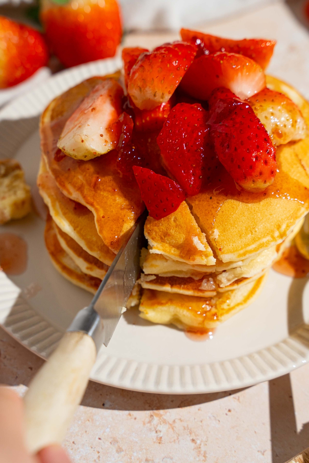 A white plate with a stack of IHOP New York cheesecake pancakes with a strawberry topping. A knife is slicing a bite from the stack. The plate is on a tan counter with strawberries and a white cloth napkin.