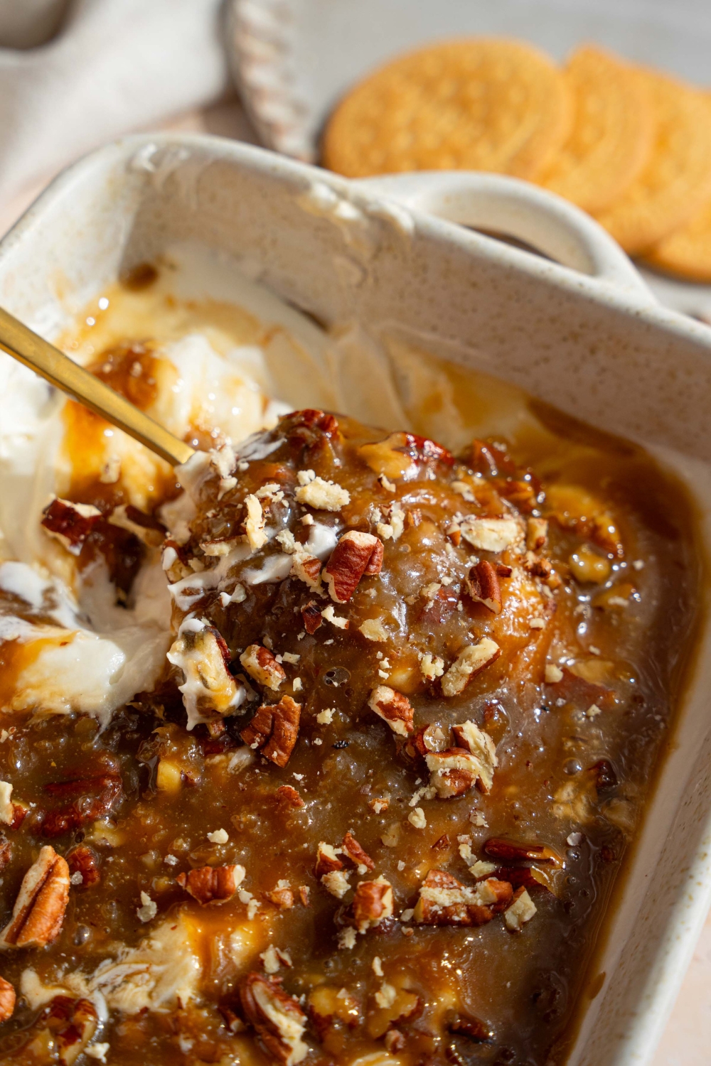 A white baking sheet with no bake cheesecake dip topped with a chilled pecan pie topping. A spoon is scooping into the dip. The dish is on a tan counter with a white cloth napkin and small plate of cookies.