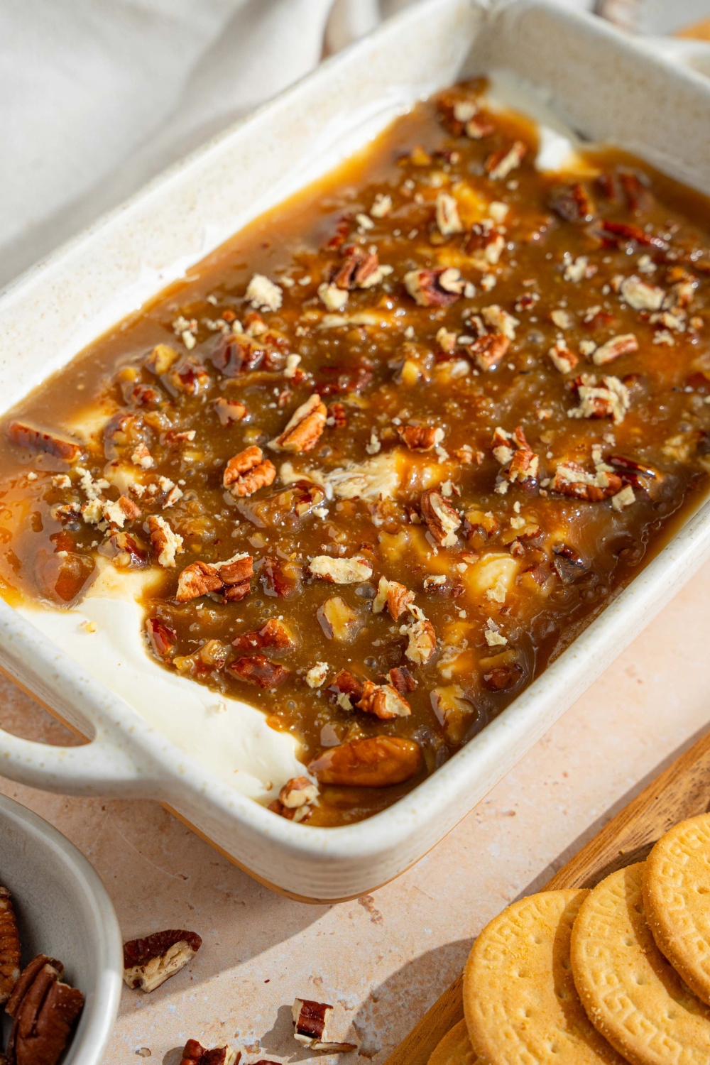 A white baking sheet with no bake cheesecake dip topped with a chilled pecan pie topping. The dish is on a tan counter with a white cloth napkin and small plate of cookies.