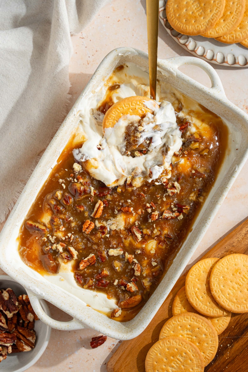 A white baking sheet with no bake cheesecake dip topped with a chilled pecan pie topping. A spoon and vanilla cookie is scooped into the dip. The dish is on a tan counter with a white cloth napkin and small plate of cookies.