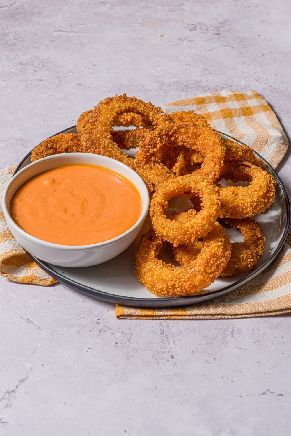 A plate of fried onion rings with a bowl of boom boom sauce. The plate is on a stone counter with a yellow striped napkin.