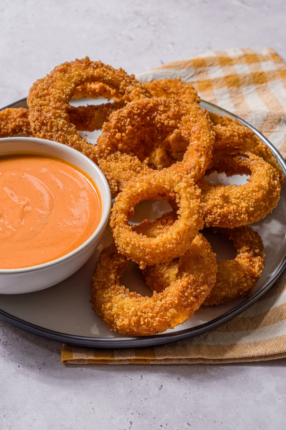 A plate of fried onion rings with a bowl of boom boom sauce. The plate is on a stone counter with a yellow striped napkin.