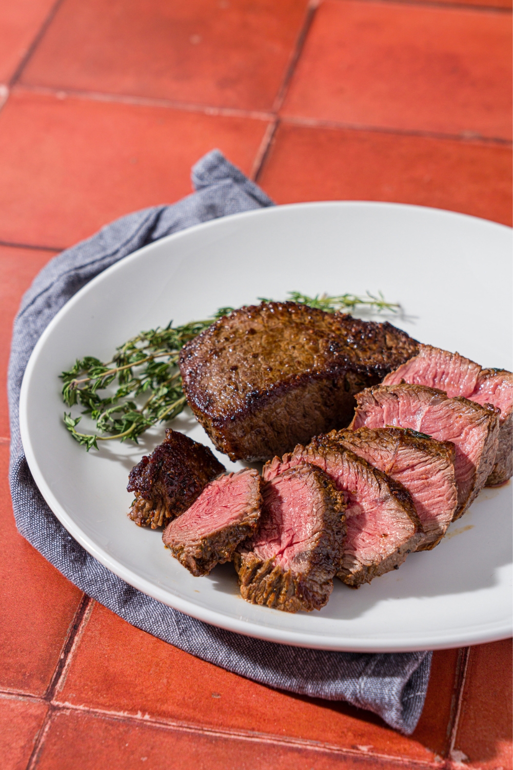 A white plate with sliced petite sirloin steak next to a whole petite sirloin and sprig of thyme. The plate is on a blue linen napkin on red tiled counter.