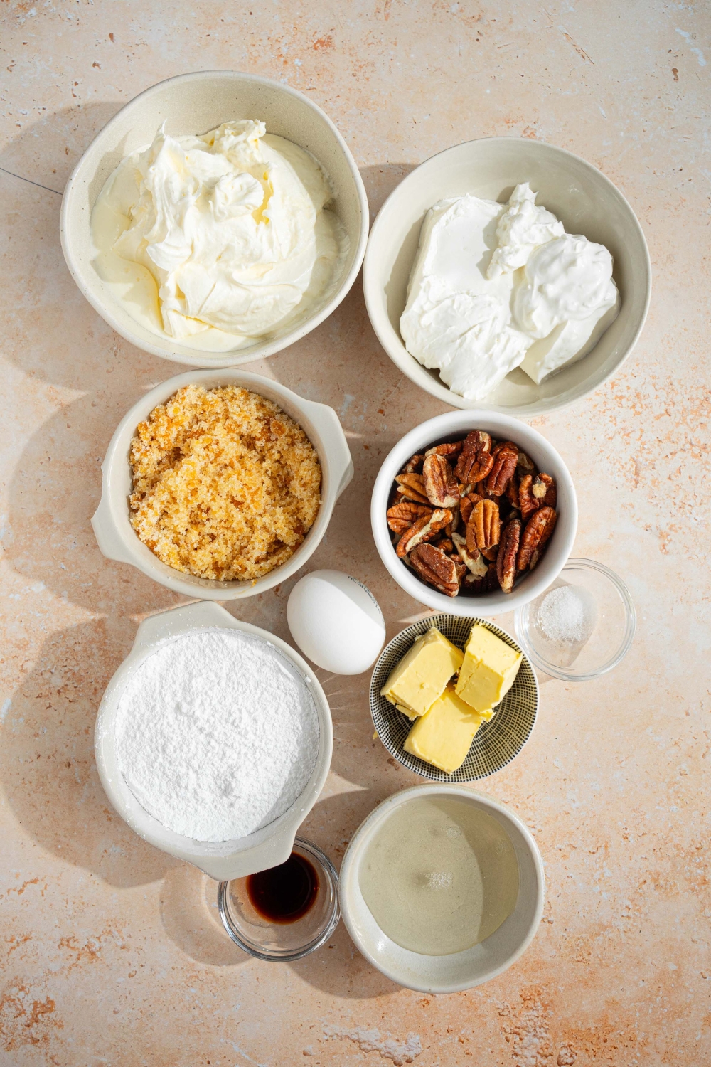 An overhead shot of several bowls containing ingredients to make no bake cheesecake with pecan pie topping including cream cheese, whipped cream, egg, butter, pecans, corn syrup, brown sugar, powdered sugar, and vanilla.
