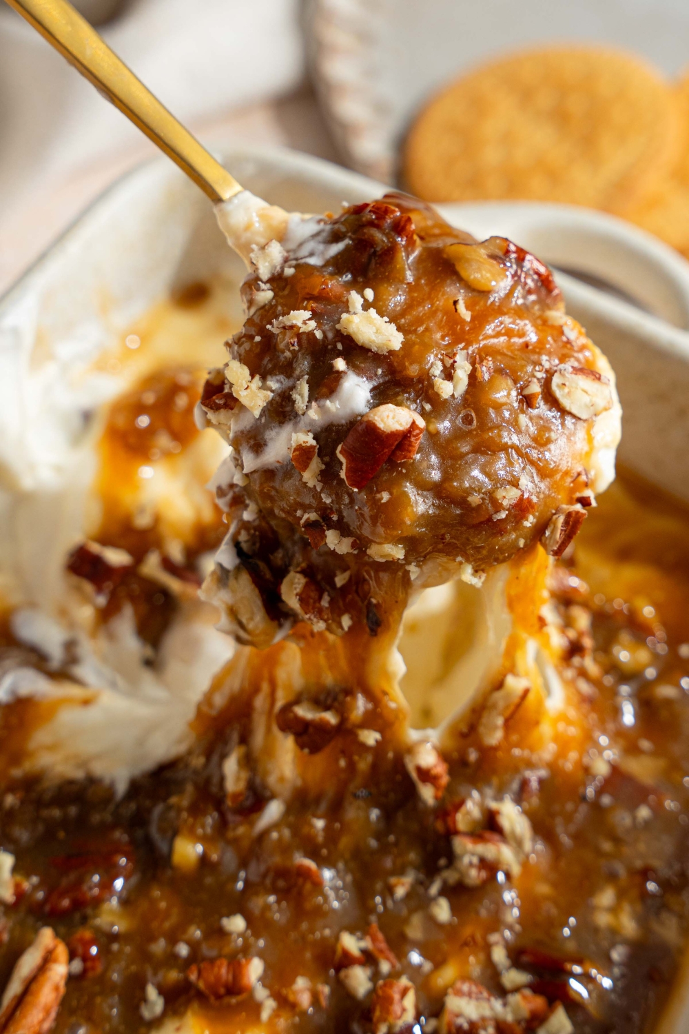 A close up of a spoon dipped into a white baking dish with no bake cheesecake dip topped with pecan pie topping. The dish is on a tan counter with a small plate of vanilla cookies.
