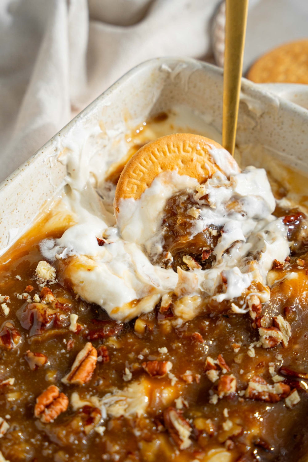 A white baking sheet with no bake cheesecake dip topped with a chilled pecan pie topping. A spoon is in the dip. A vanilla cookie is dipped into the dish. The dish is on a tan counter with a white cloth napkin and small plate of cookies.