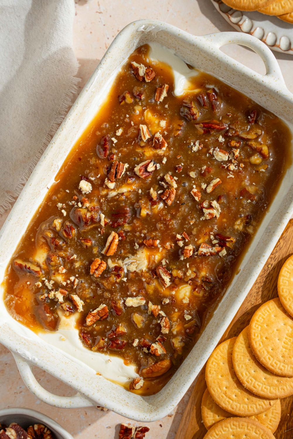 A white baking sheet with no bake cheesecake dip topped with a chilled pecan pie topping. The dish is on a tan counter with a white cloth napkin and small plate of cookies.