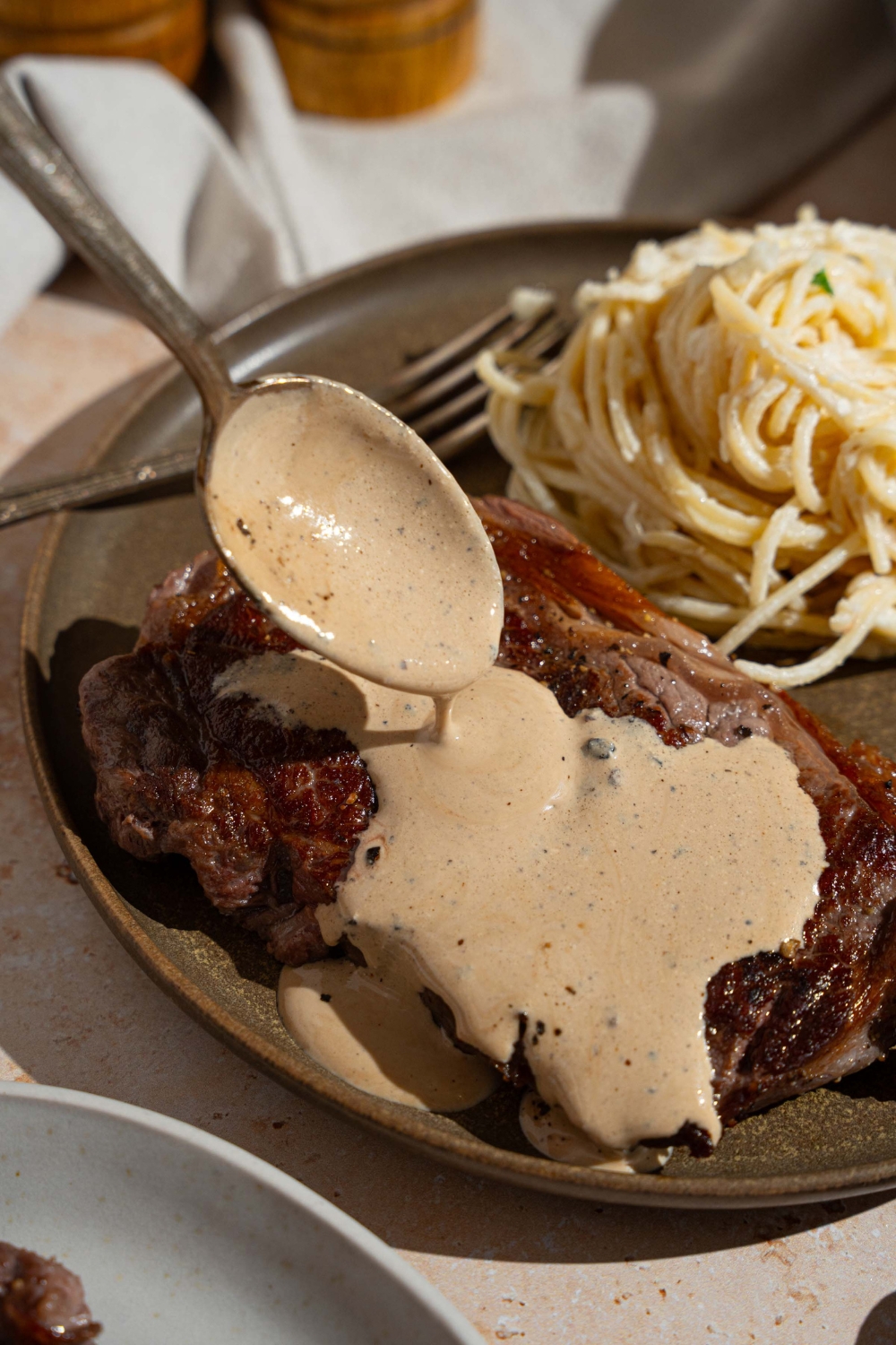A spoon pouring peppercorn sauce over cooked steak on a plate with a side of spaghetti. The plate is on a tan counter.