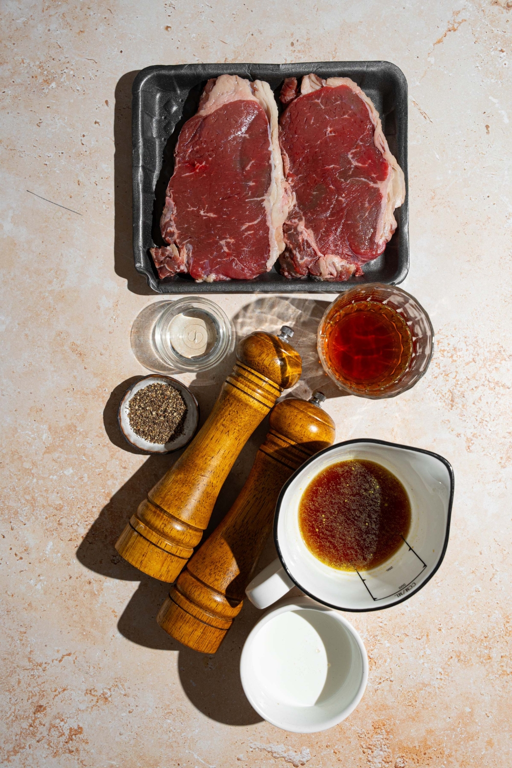 An overhead shot of several bowls in various sizes containing ingredients to make peppercorn sauce including steaks, brandy, beef broth, heavy cream, peppercorns, and a salt and pepper mill.