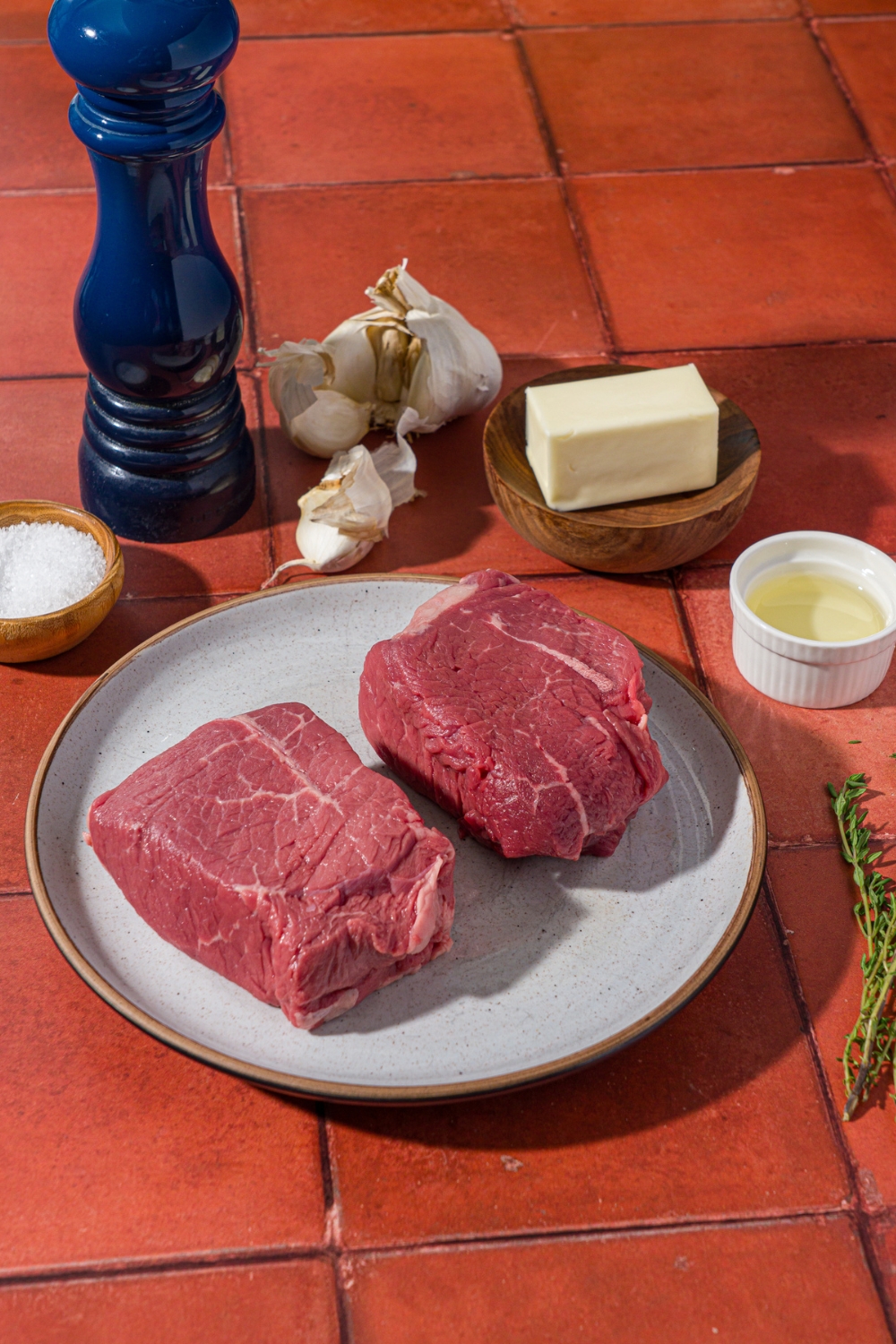 A red tiled counter with ingredients to make petite sirloin steak including two petite sirloin steaks, butter, oil, garlic, salt, thyme, and a pepper mill.