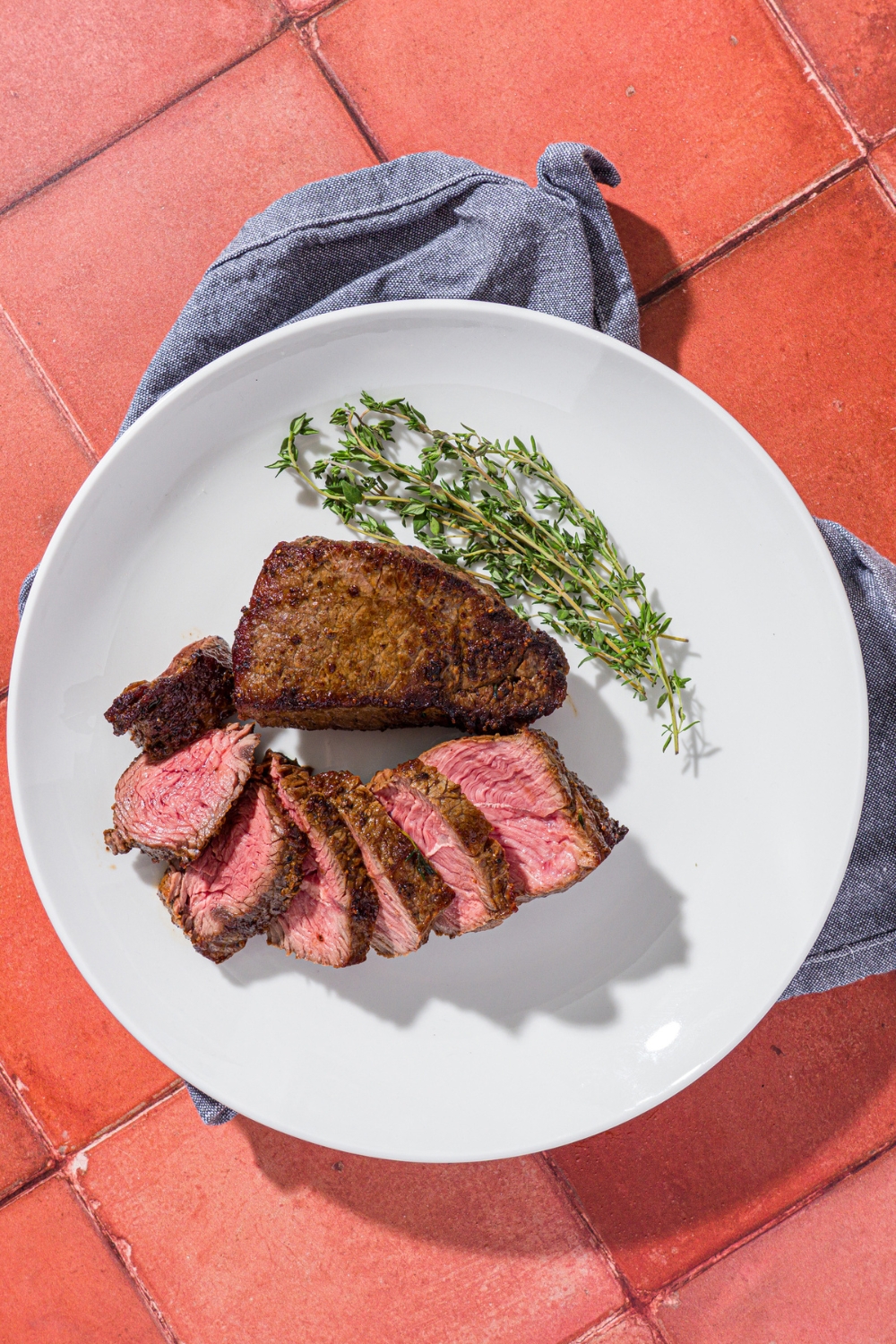 A white plate with sliced petite sirloin steak next to a whole petite sirloin and sprig of thyme. The plate is on blue linen napkin a red tiled counter.