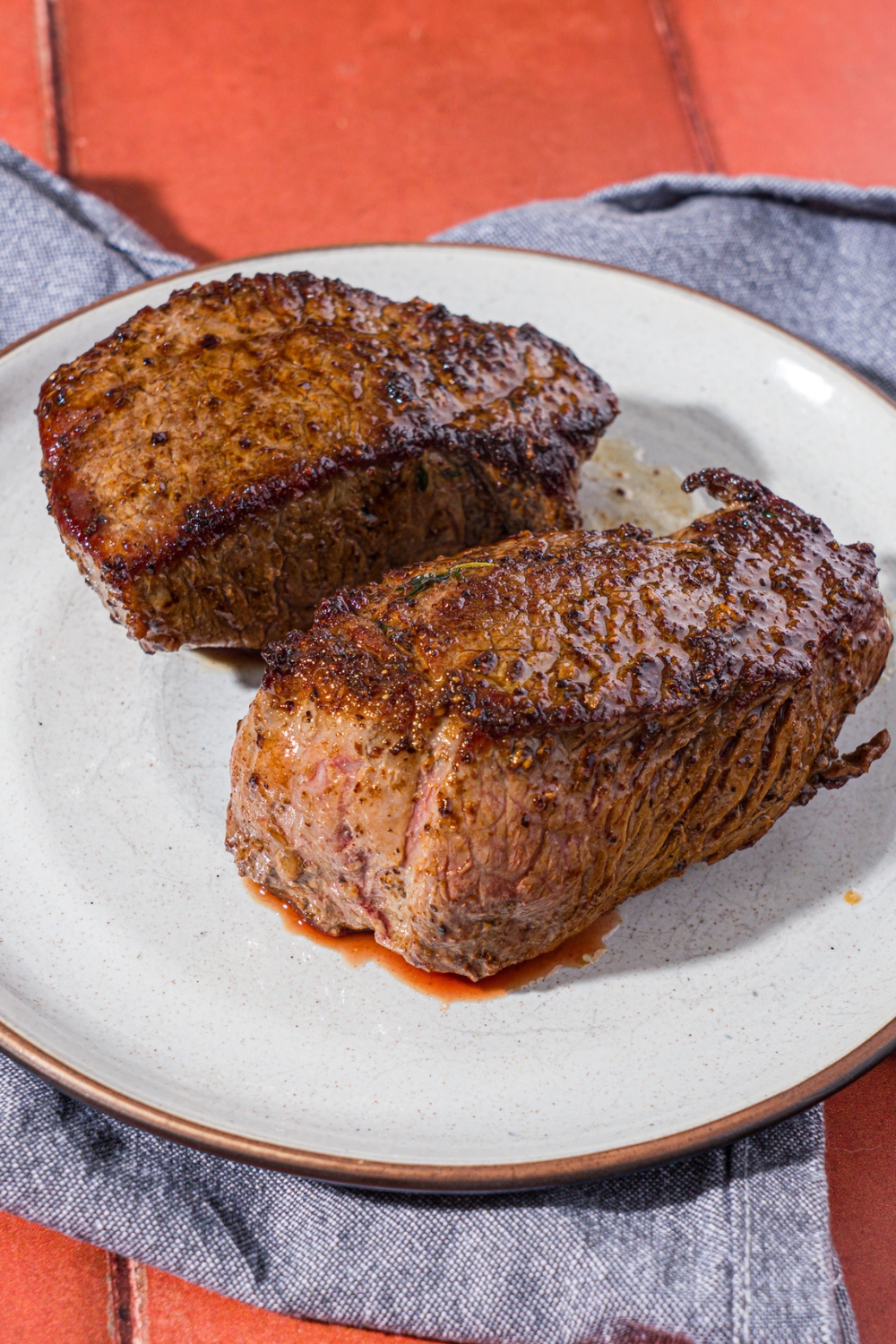 Two whole cooked petite sirloin steaks on a white plate. The plate is on a blue linen napkin on a red tiled counter.