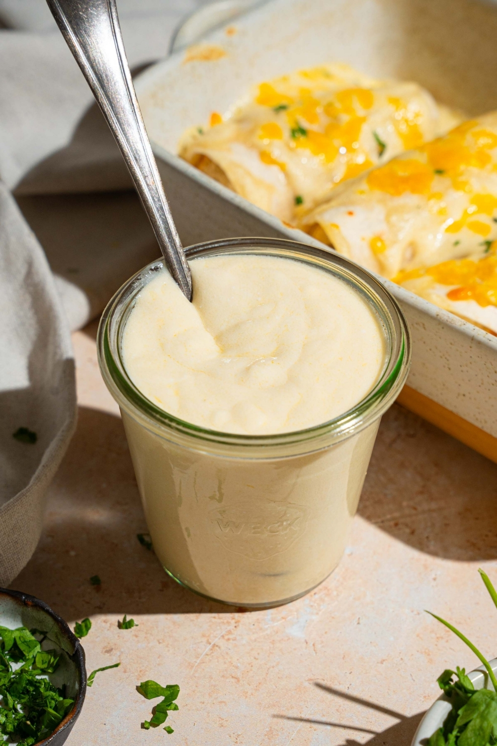 A jar of sour cream sauce for enchiladas with a spoon in the jar. The jar is on a white counter with a baking dish of baked enchiladas and small bowls of garnishes.