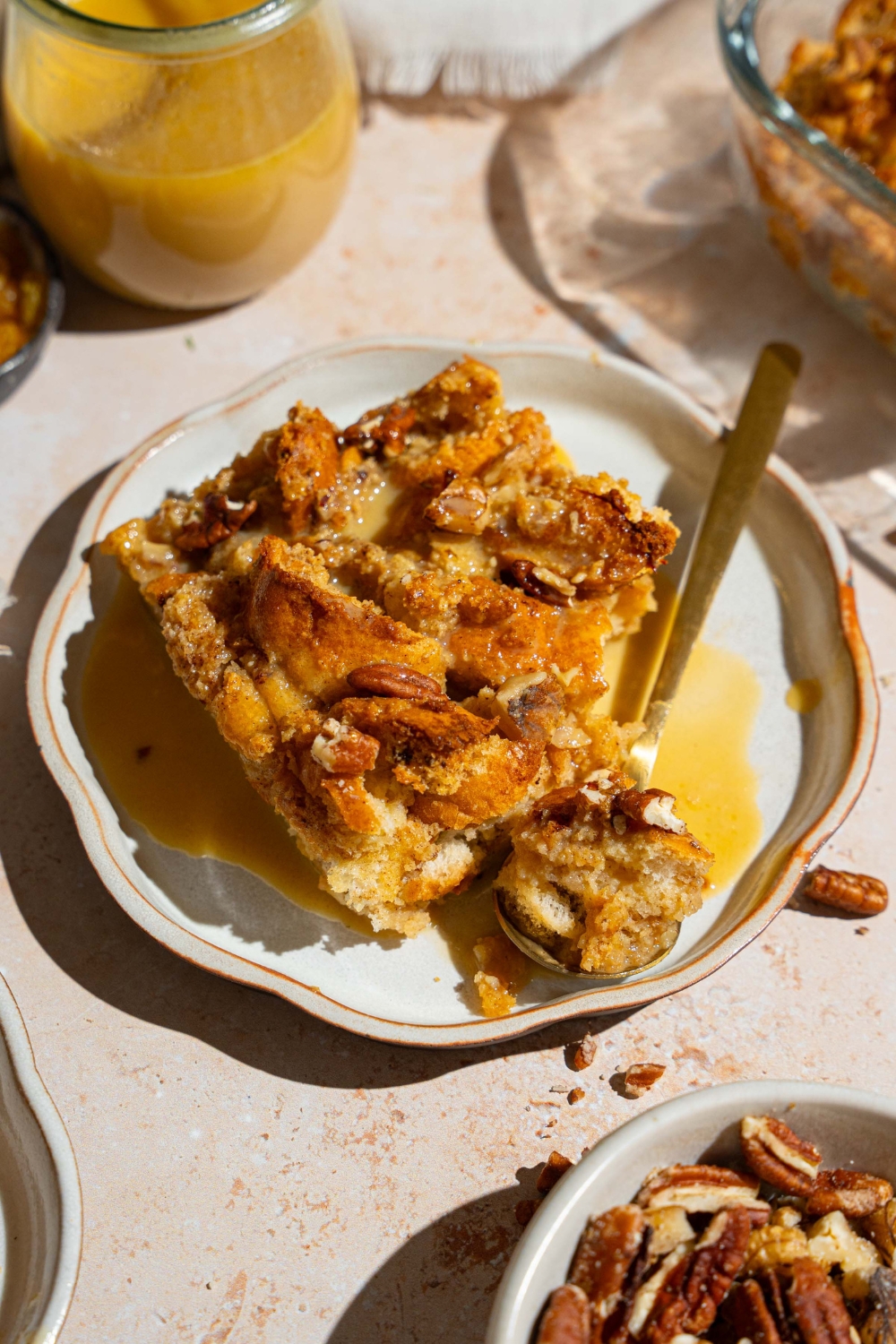 A white plate with a slice of bread pudding topped with rum sauce with a spoon with a bite of bread pudding. The plate is on a tan counter with a jar of rum sauce and small bowl of pecans.