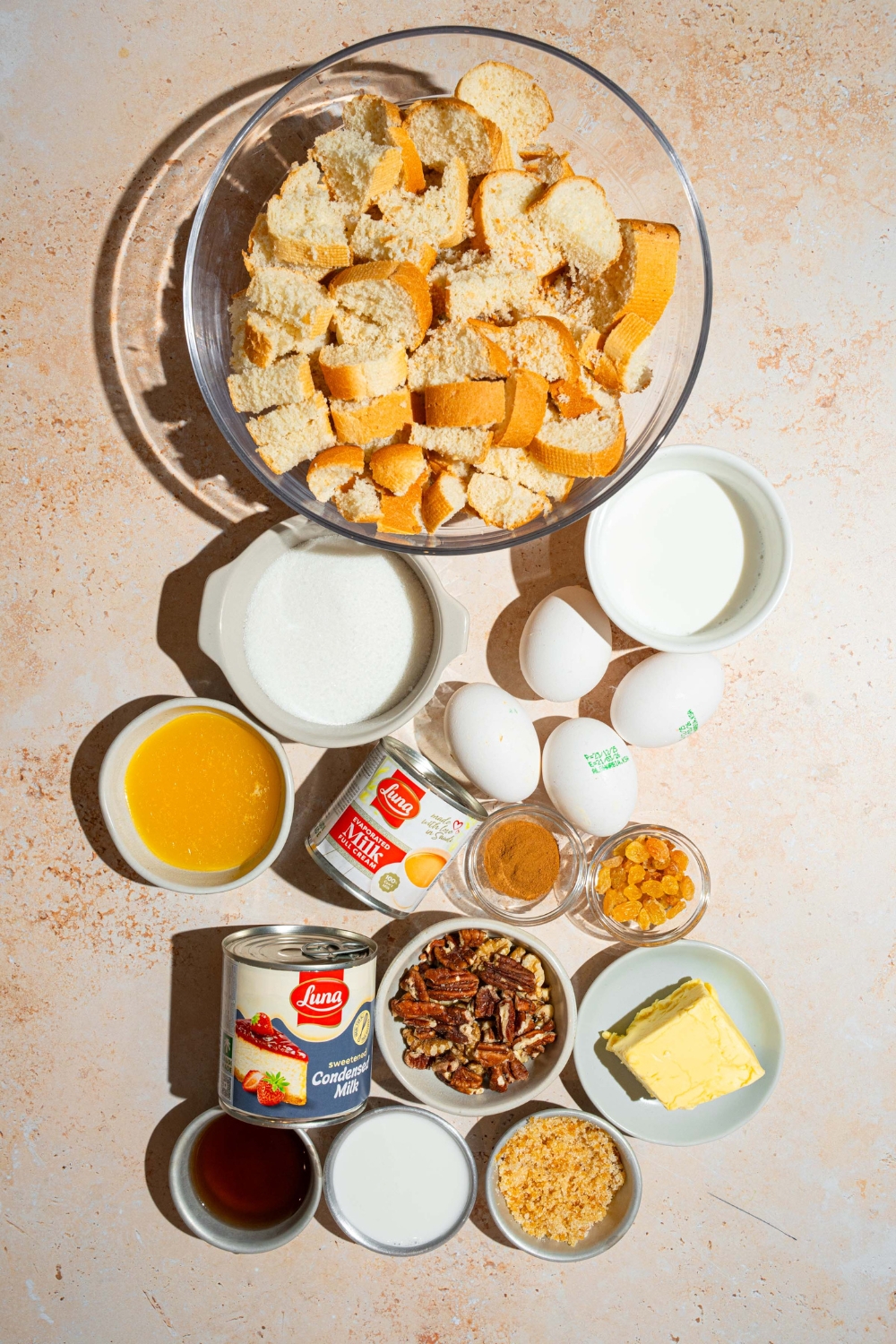 An overhead shot of several bowls in various sizes containing ingredients to make rum sauce for bread pudding including cubed bread, condensed milk, cream, butter, rum, brown sugar, rum extract, eggs, and more.