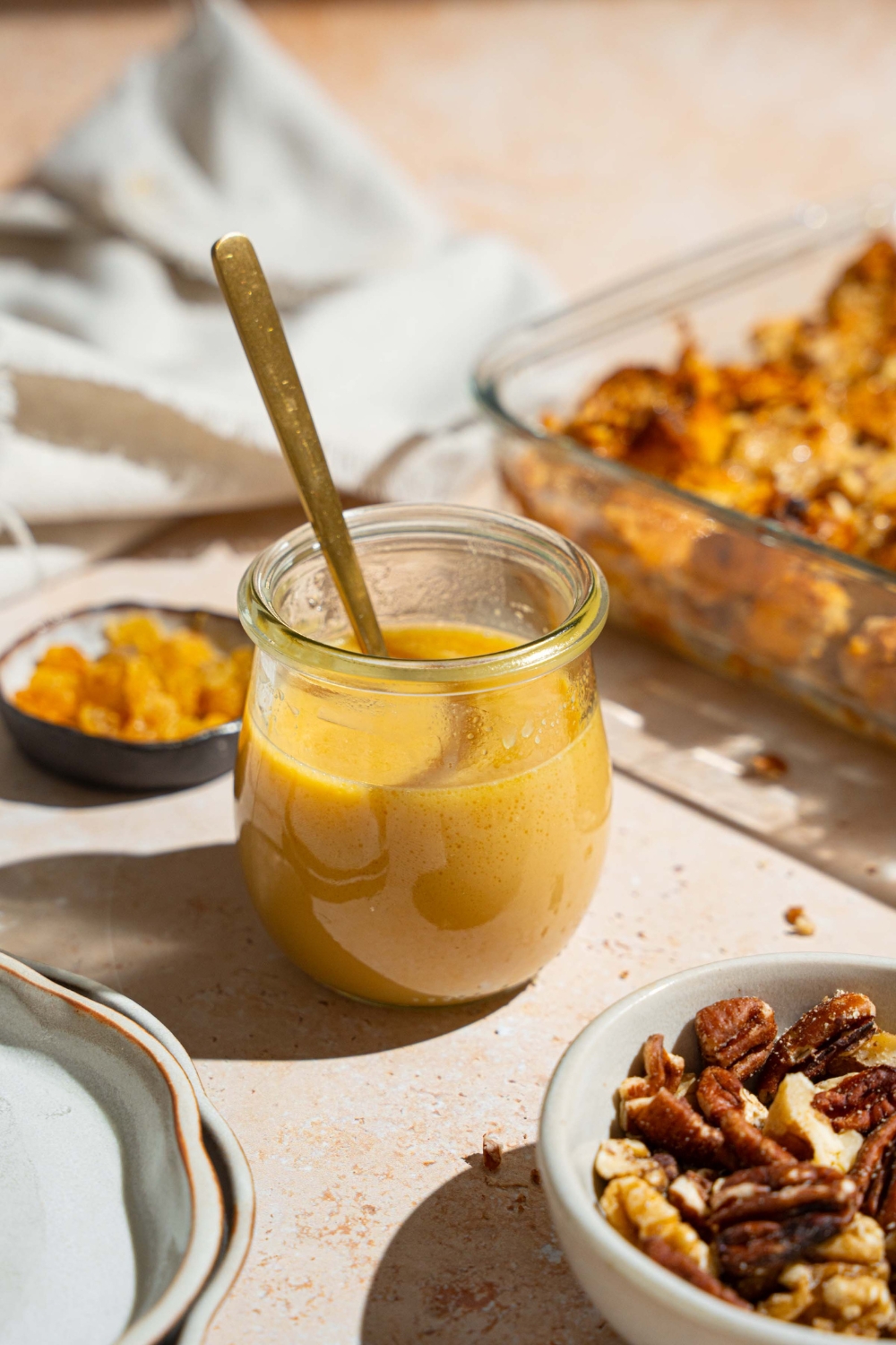 A glass jar with rum sauce for bread pudding with a spoon in the jar. The jar is on a tan counter with a plate of bread pudding and small bowl of golden raisins and bowl of pecans.