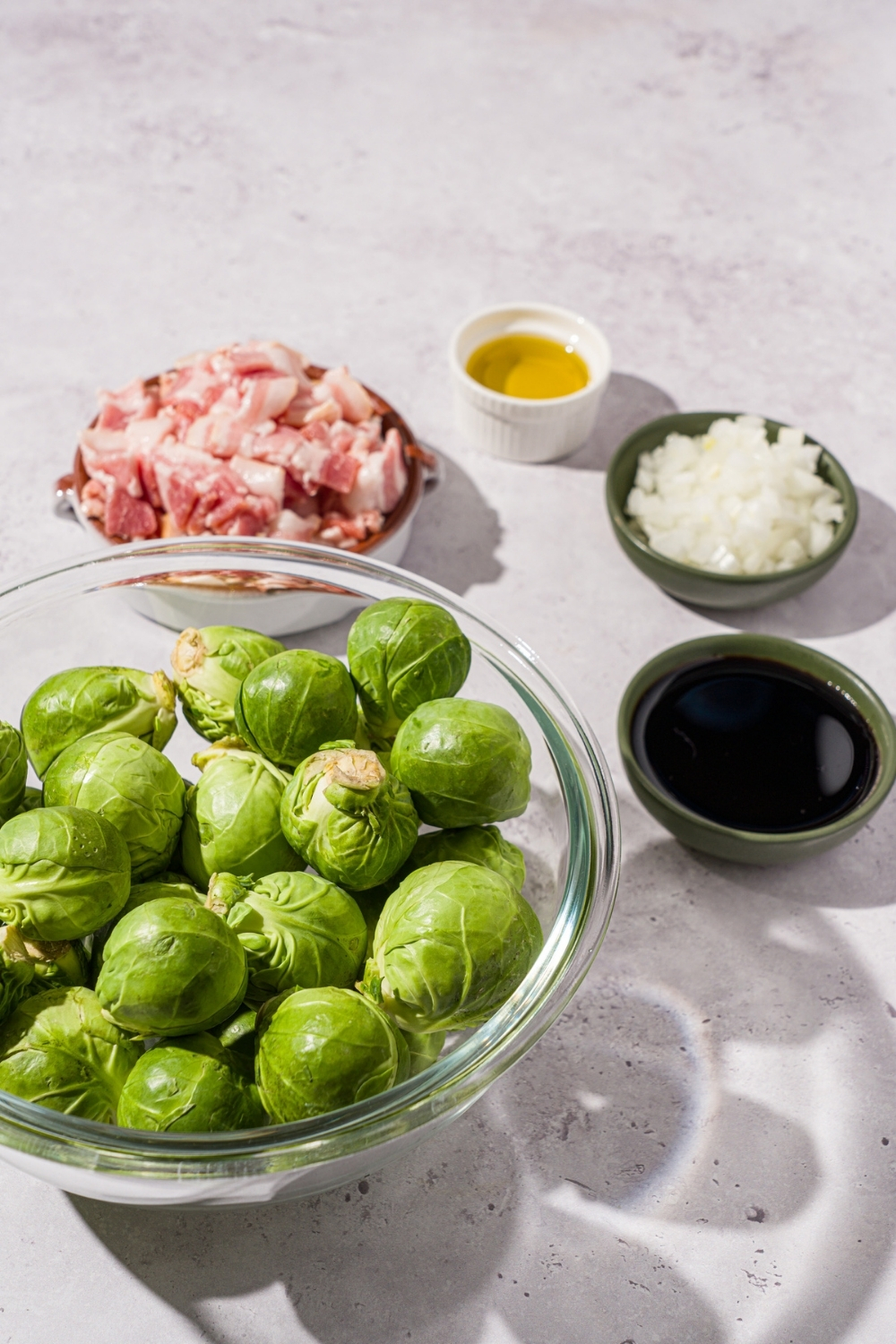 Several bowls in various sizes containing ingredients to make shaved brussel sprouts including brussel sprouts, uncooked bacon, diced onion, oil, and balsamic reduction.