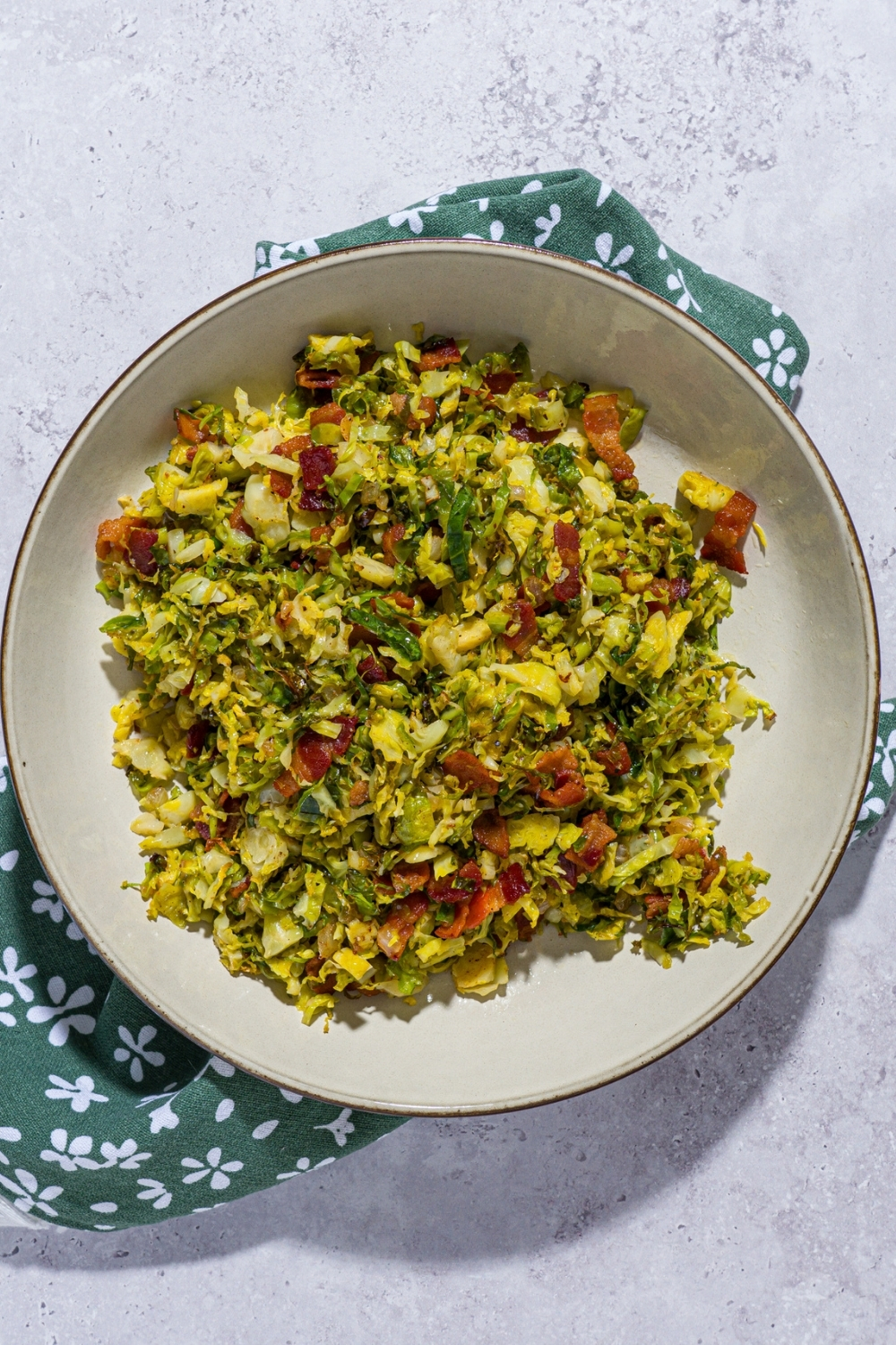 A bowl of shaved brussel sprouts salad tossed with bacon and onion and drizzled with balsamic reduction. The bowl is on a green floral napkin on a white counter.