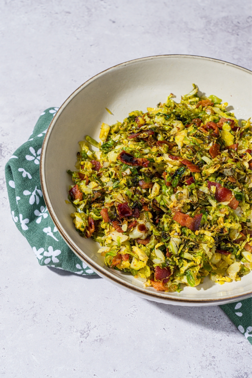 A bowl of shaved brussel sprouts salad tossed with bacon and onion and drizzled with balsamic reduction. The bowl is on a green floral napkin on a white counter.