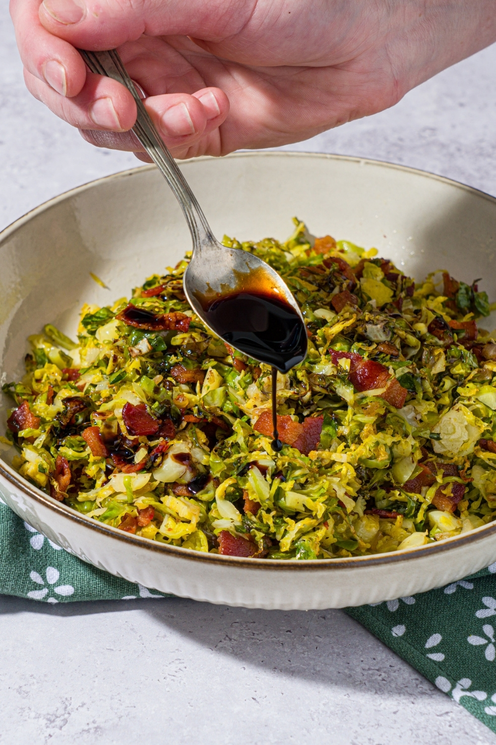 A bowl of shaved brussel sprouts salad tossed with bacon and onion. A hand is pouring balsamic reduction over the salad with a spoon. The bowl is on a green floral napkin on a white counter.
