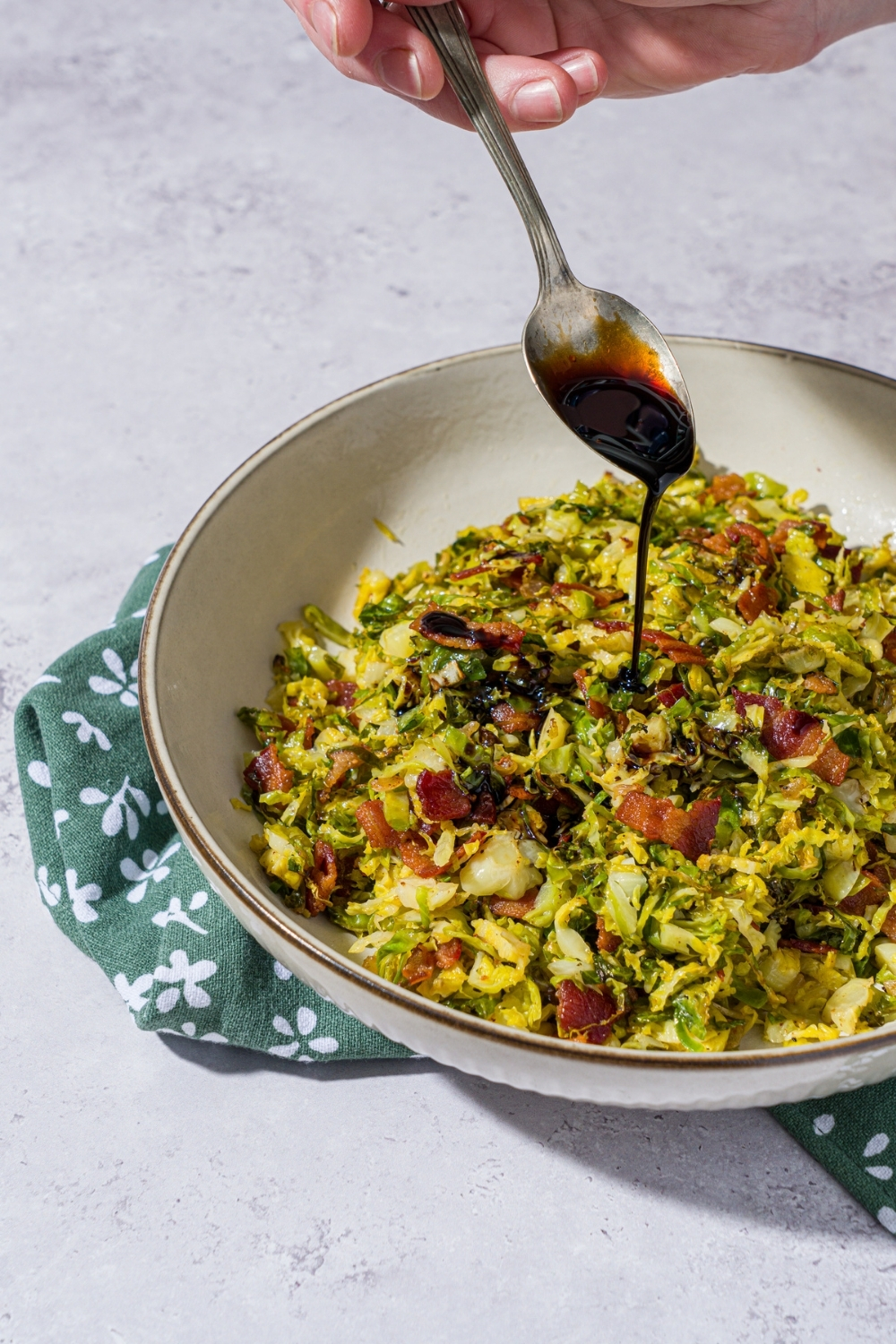 A bowl of shaved brussel sprouts salad tossed with bacon and onion. A hand is pouring balsamic reduction over the salad with a spoon. The bowl is on a green floral napkin on a white counter.