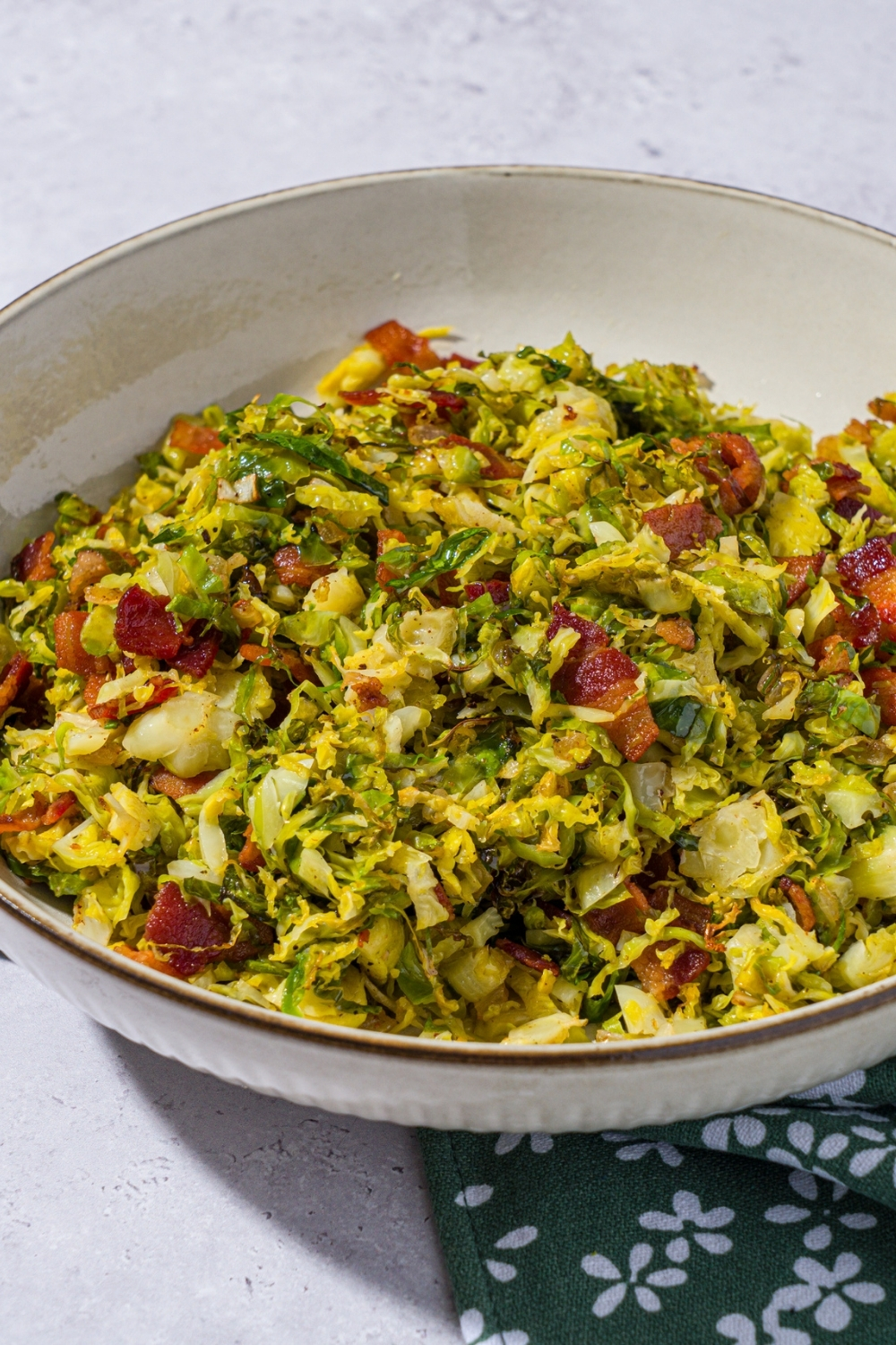 A bowl of shaved brussel sprouts salad tossed with bacon and onion and drizzled with balsamic reduction. The bowl is on a green floral napkin on a white counter.