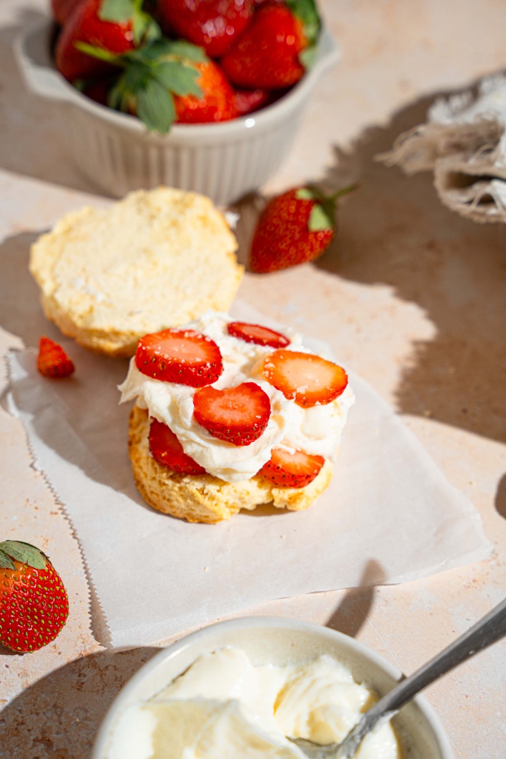 An open strawberry shortcake with layers of whipped cream and sliced strawberries on a shortcake biscuit. The shortcake is on a piece of parchment paper on a tan counter with a bowl of strawberries and bowl of whipped cream.